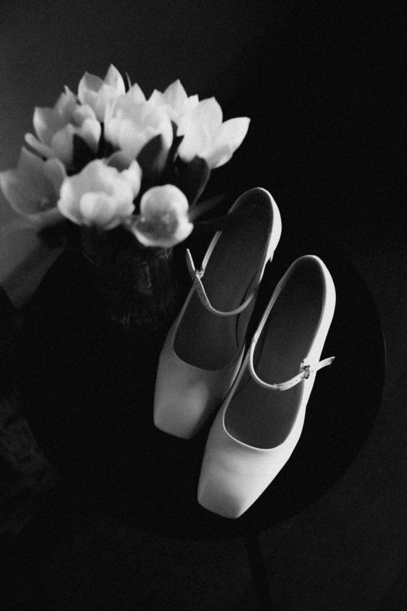 Black and white flat lay of white Mary Jane shoes beside a vase of tulips on a dark table.