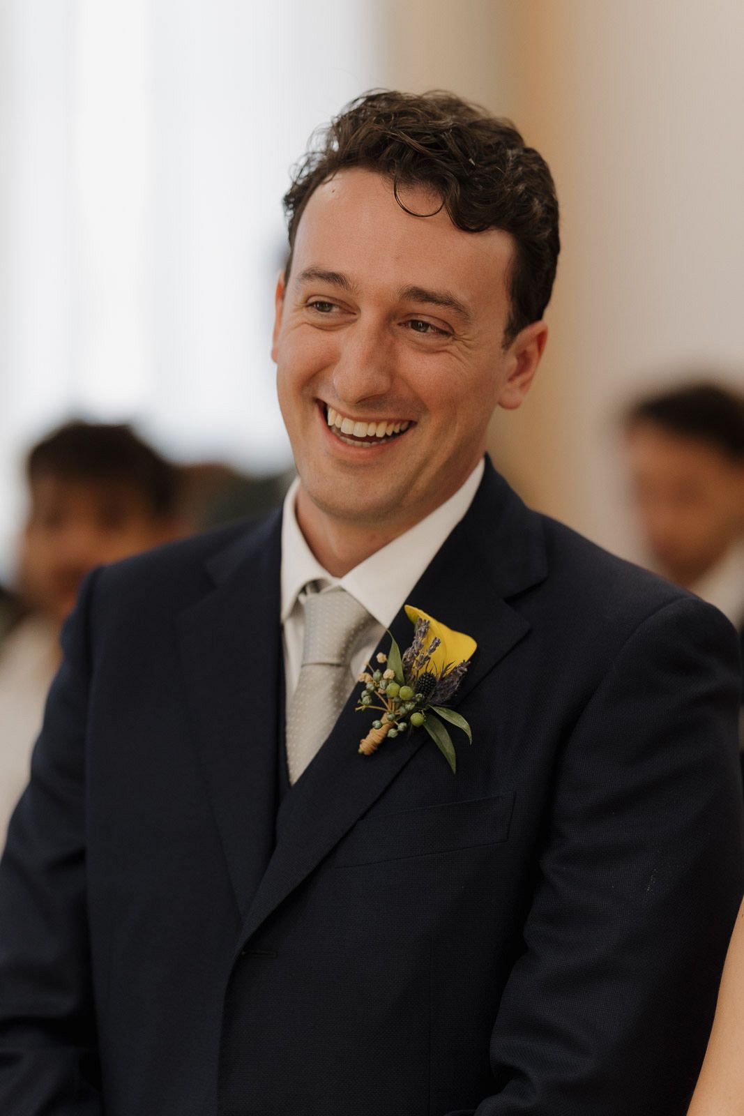 A groomsman wearing a black suit with yellow boutonniere smiles joyfully at a wedding celebration.
