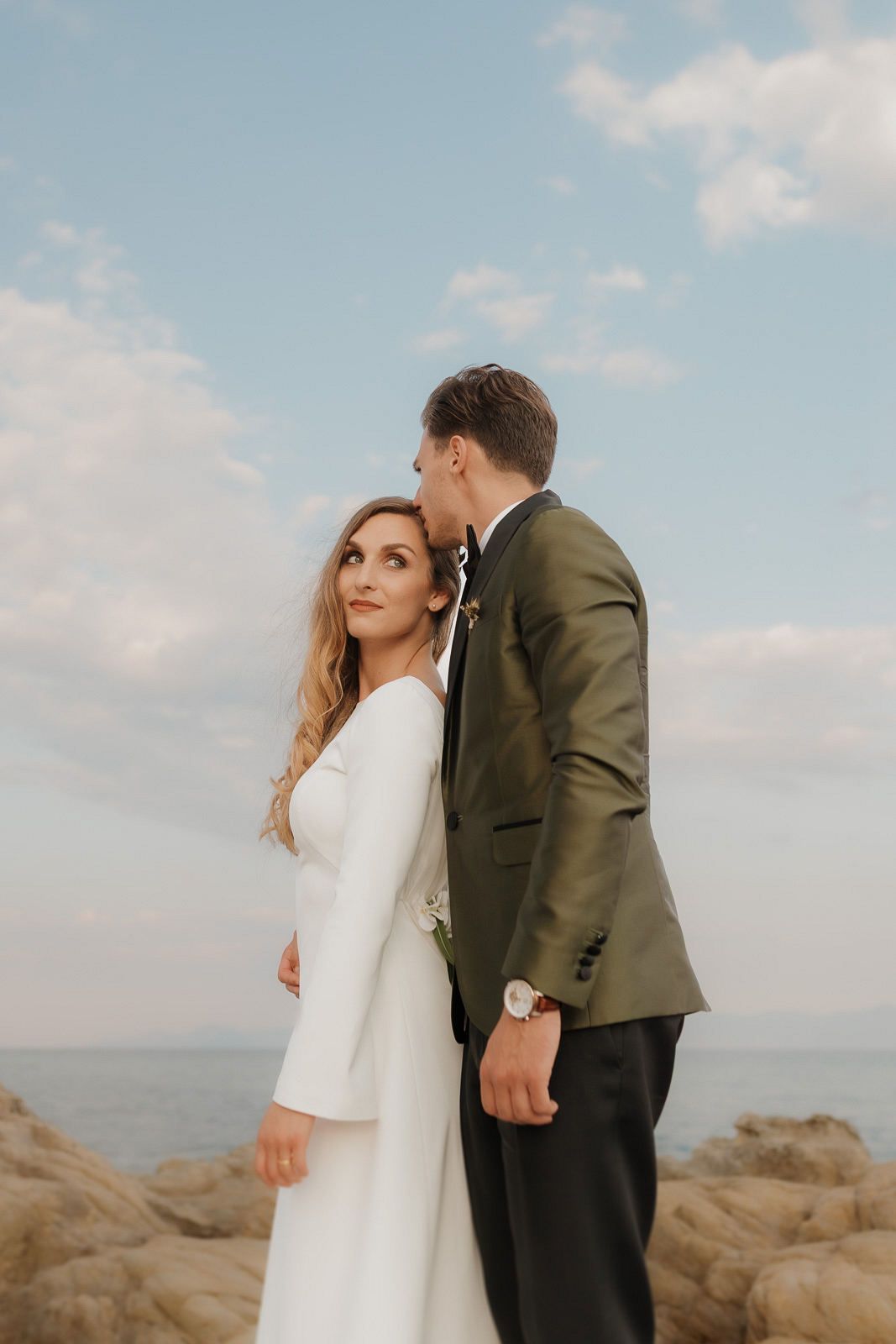 A romantic couple in wedding attire shares intimate moments along a rocky coastal shoreline at sunset.