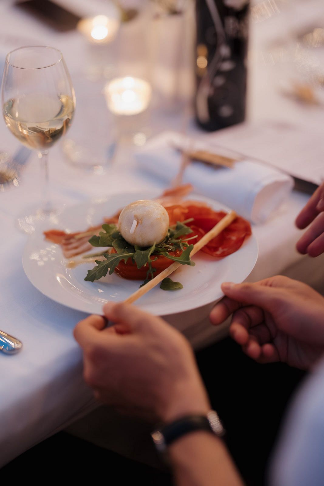 Hands holding utensils over elegant place settings with wine glasses and plates at a formal dining table.
