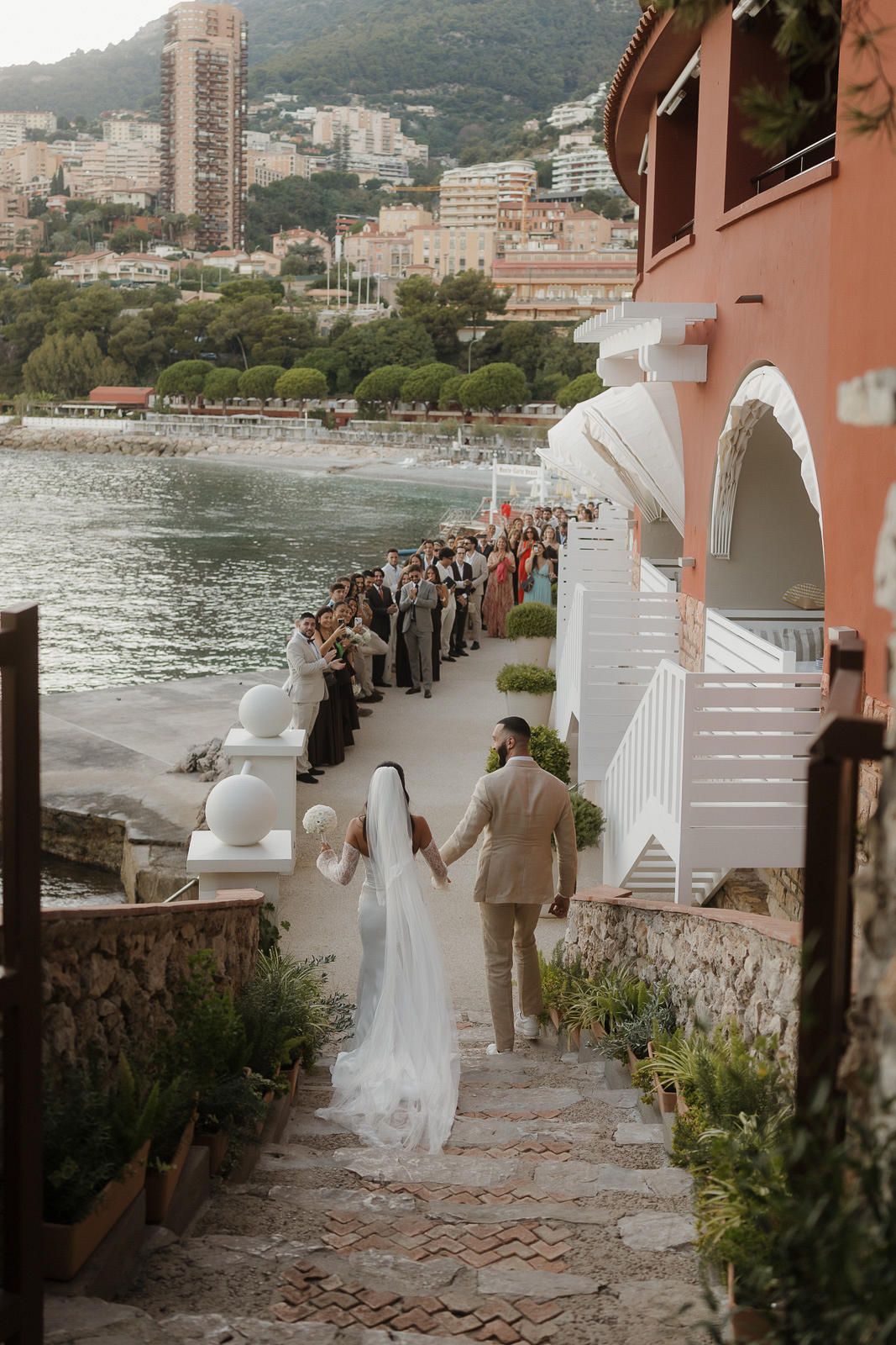 A wedding couple descends stone steps along a Mediterranean coastal villa with guests watching from a seaside walkway.
