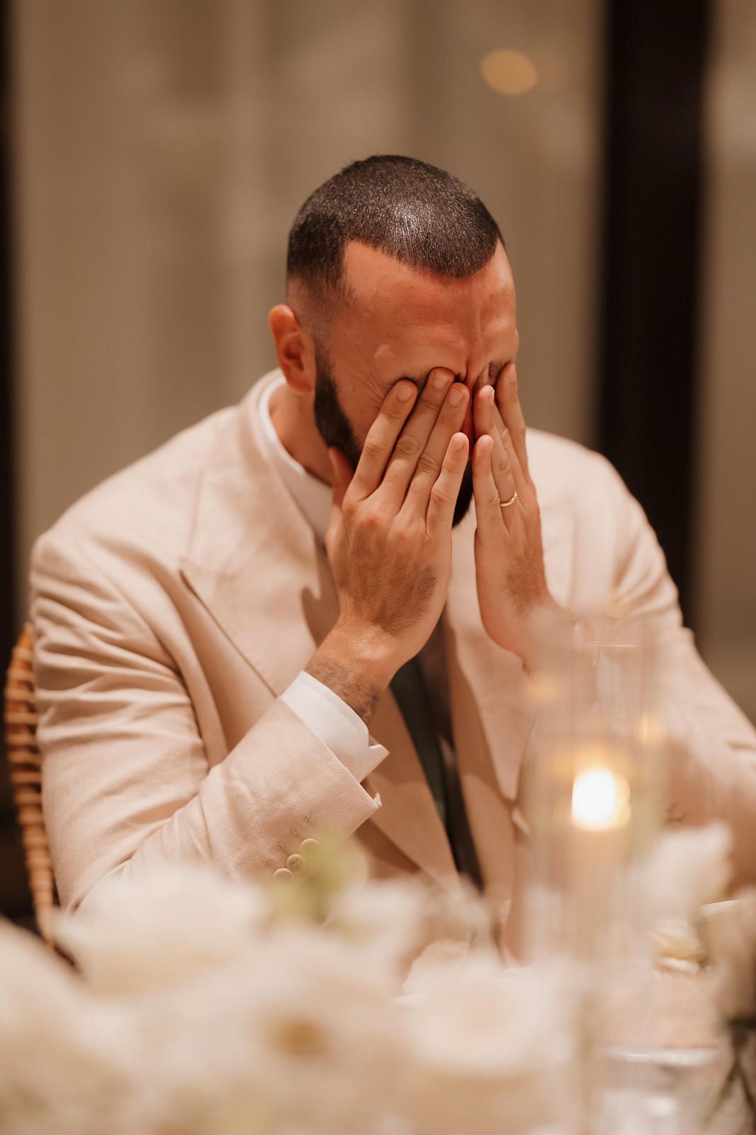 A person in a white suit jacket sits at a candlelit table with their face covered by their hands.