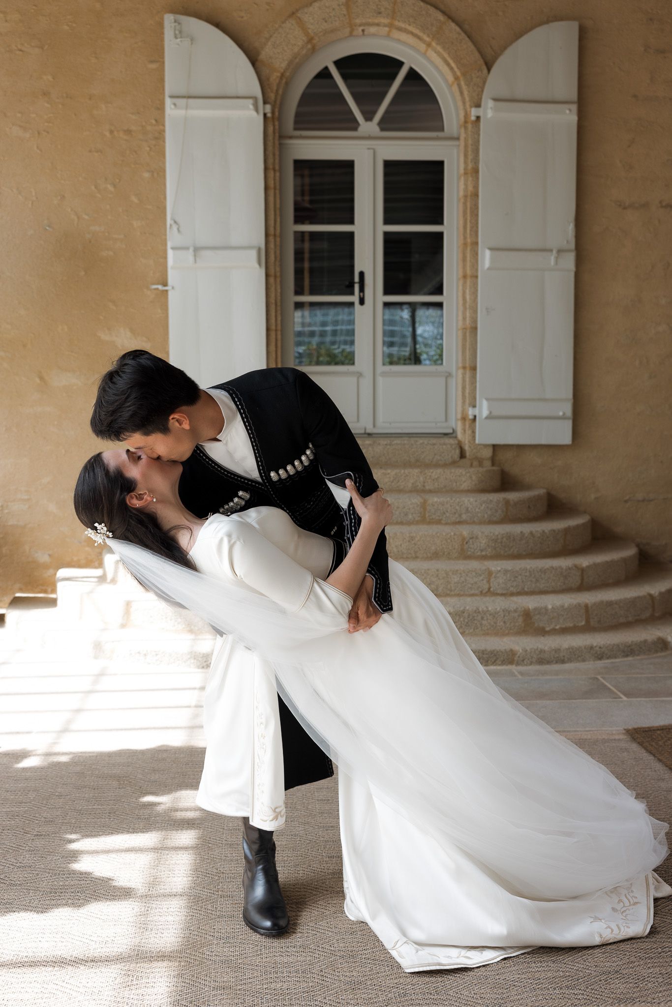 Bride and groom share a romantic dip kiss in front of a rustic stone building with arched windows and white shutters.