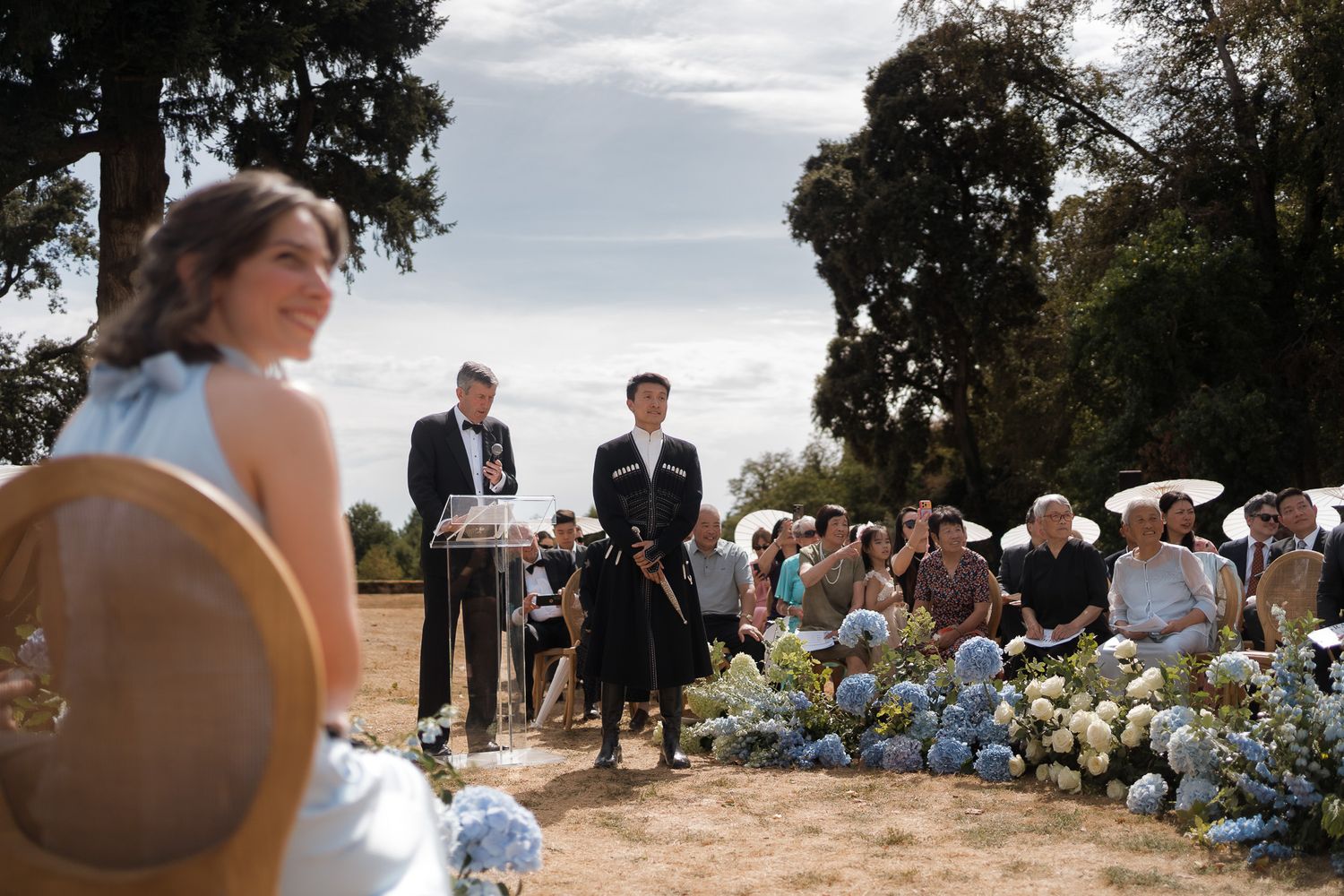 Outdoor wedding ceremony with guests seated amid blue hydrangeas, bride in foreground, officiant and groom at altar.