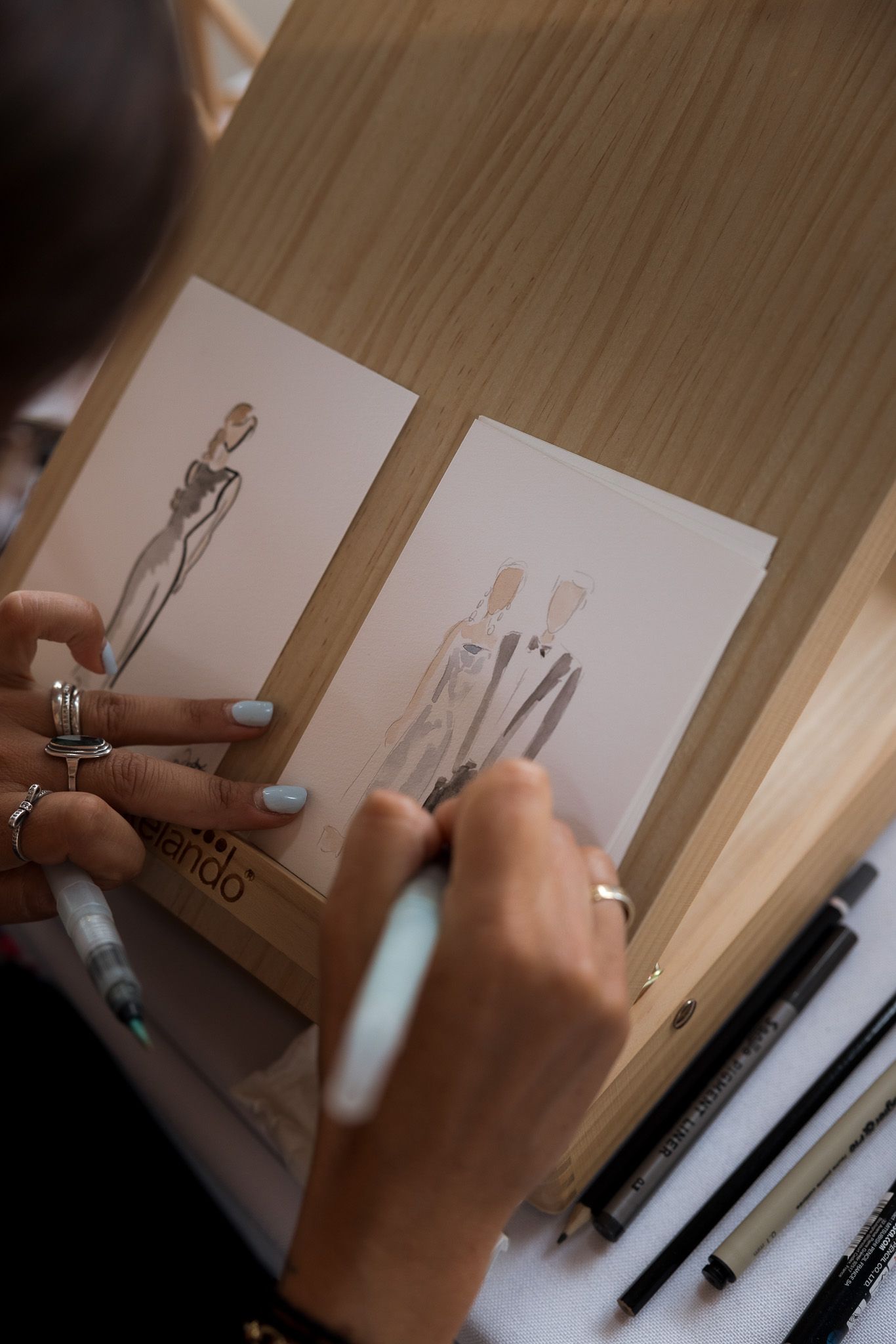 Fashion designer sketching elegant figure illustrations on paper cards propped on a wooden easel with art supplies nearby.