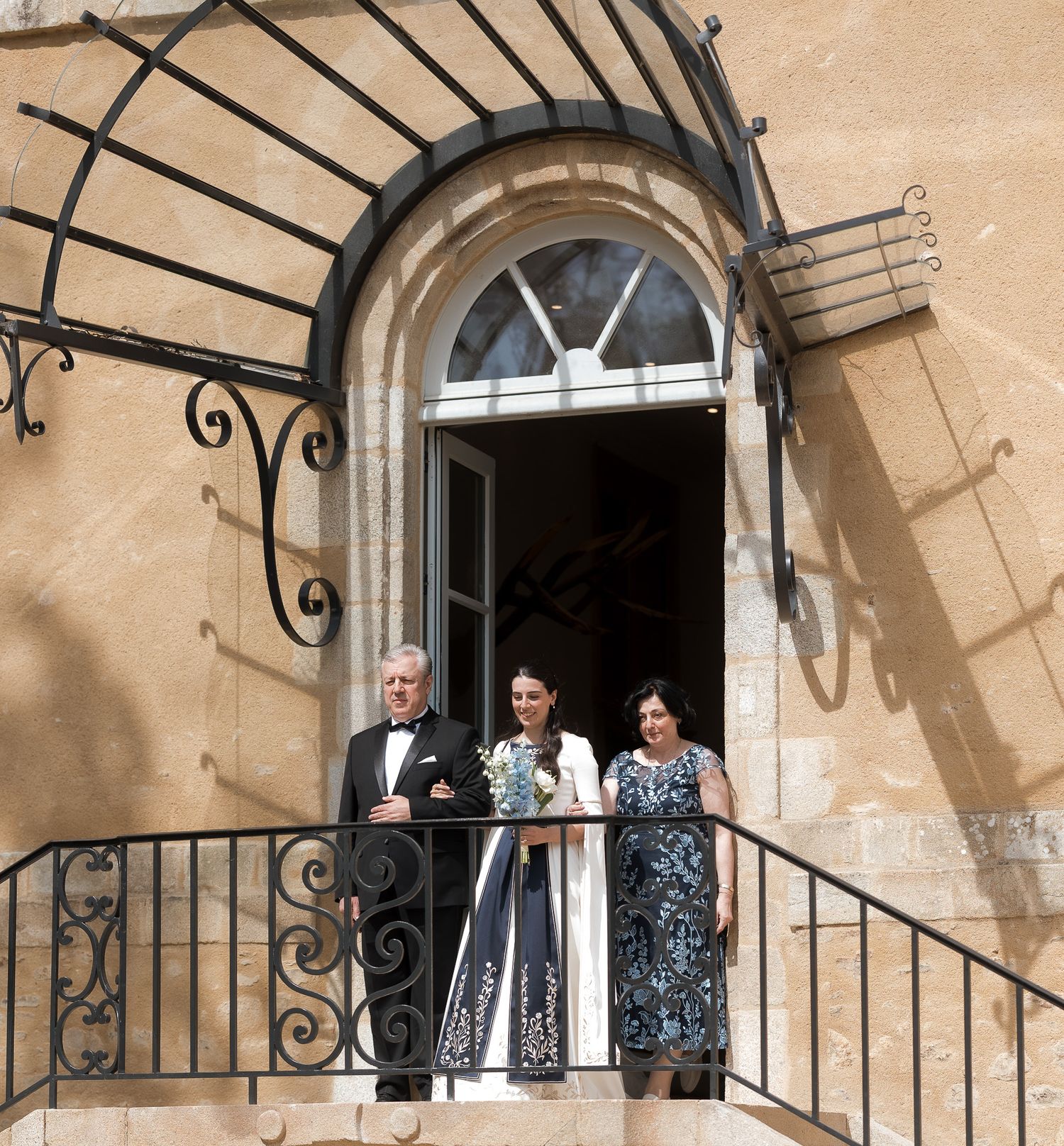 Three elegantly dressed people stand on an ornate wrought-iron balcony of a grand stone building.