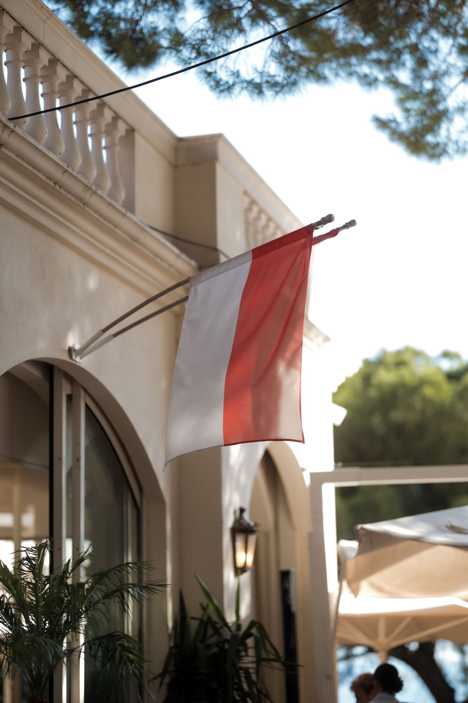 Red and white flag mounted on elegant cream building facade with arched window and lush greenery.
