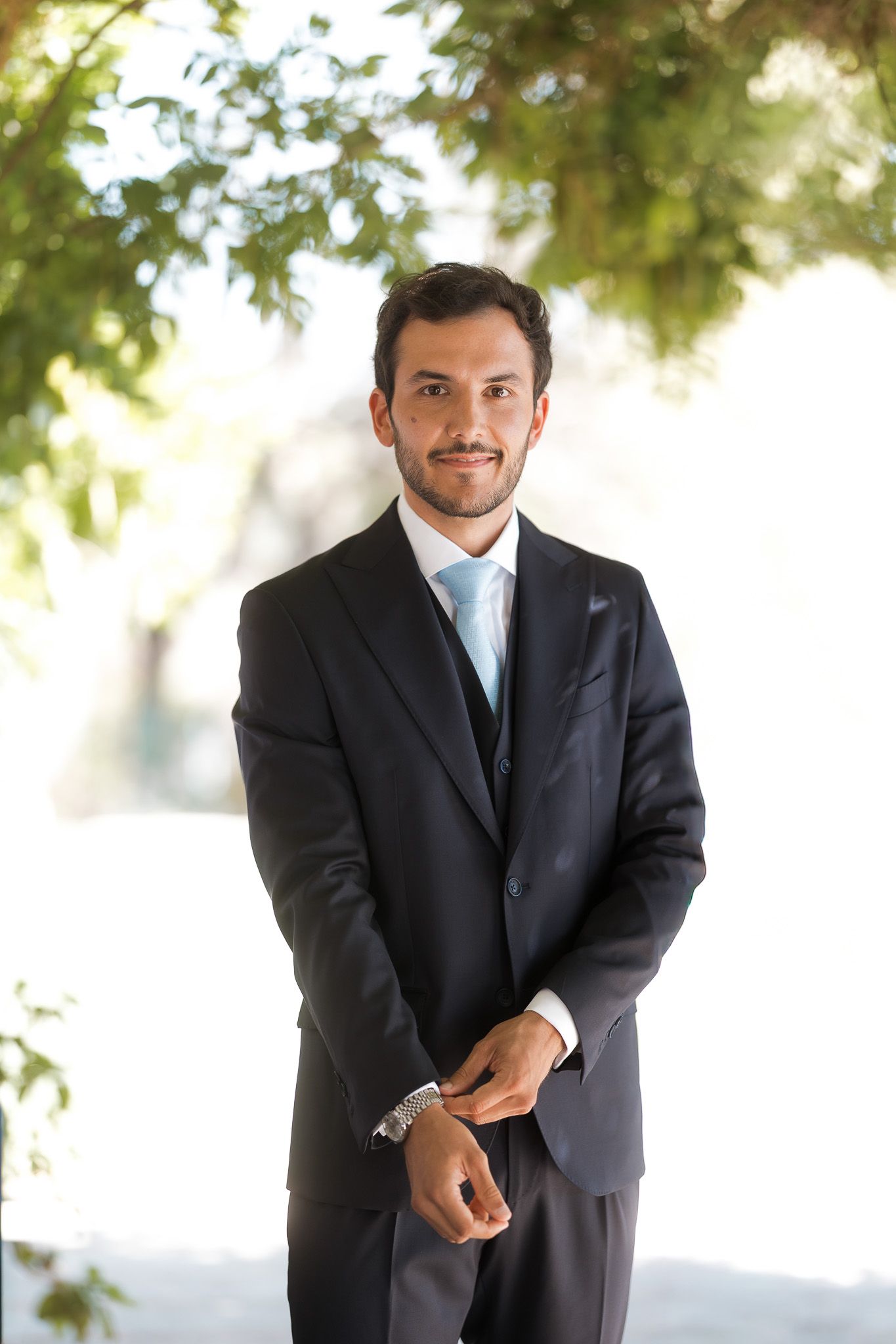 Young man in dark suit with light blue tie smiling outdoors under green trees.
