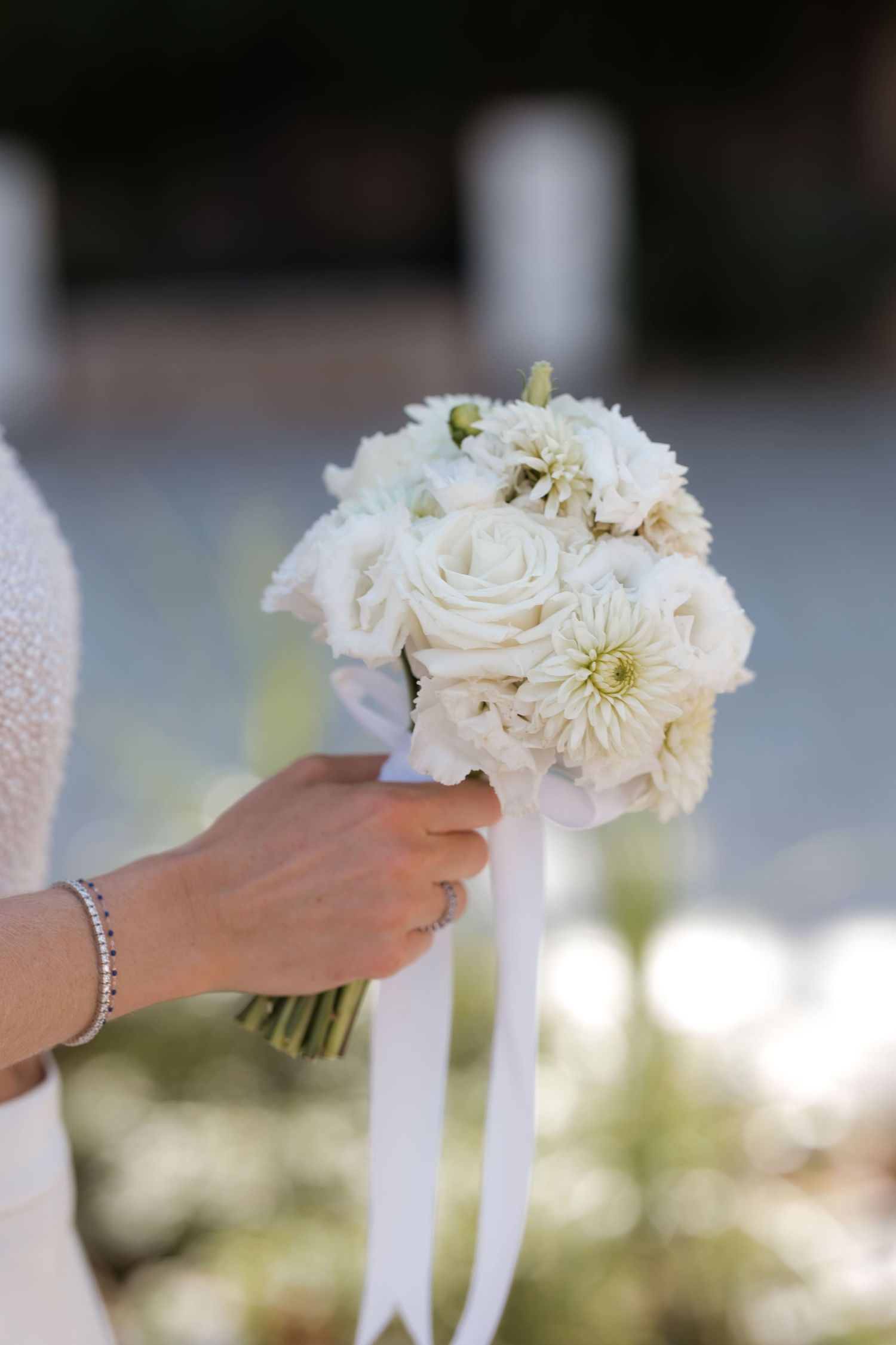 Bride holding a white wedding bouquet with roses and dahlias, tied with a satin ribbon.