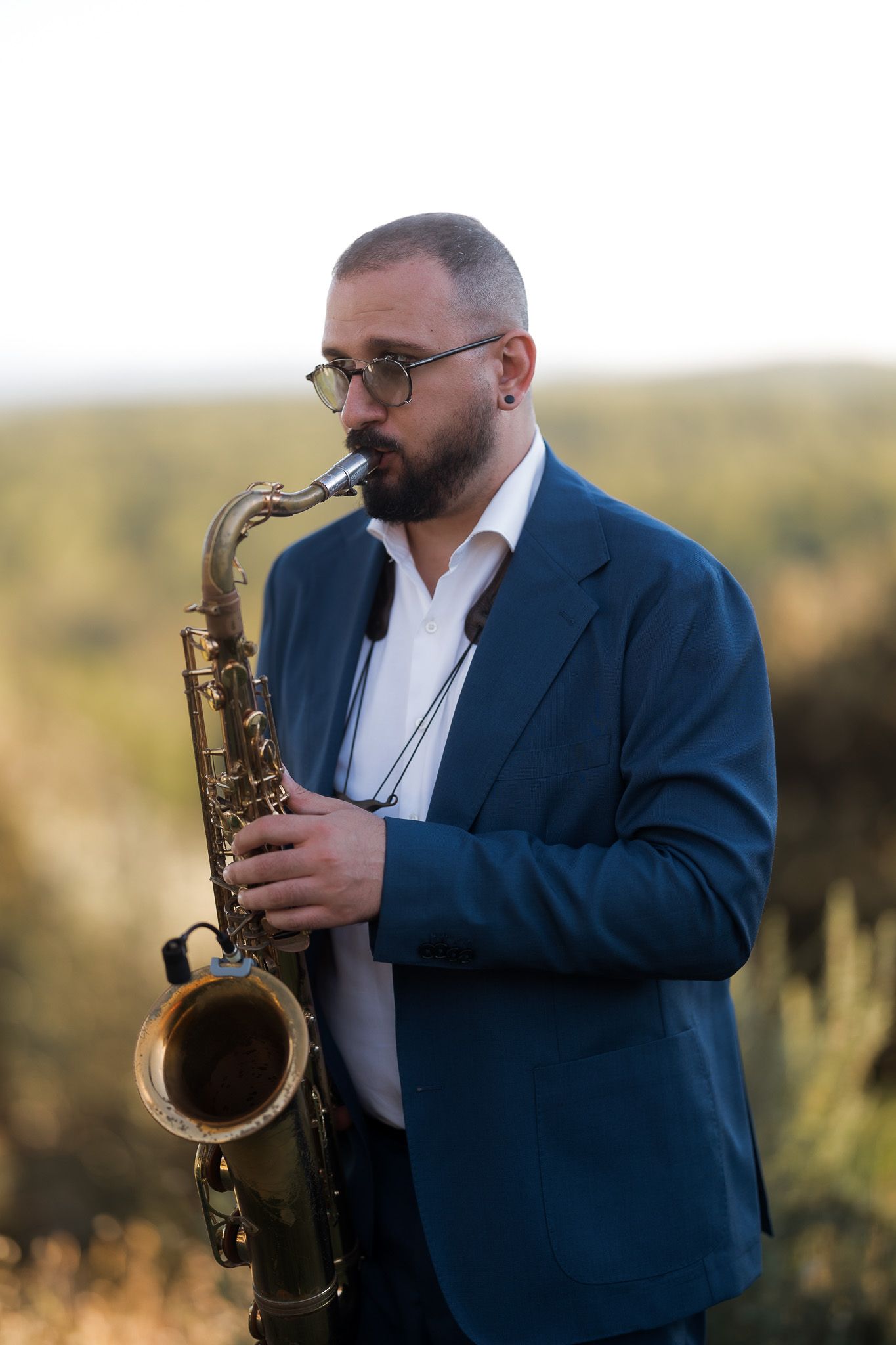 A bearded man in a blue blazer plays a gold saxophone outdoors, wearing glasses and a white shirt.
