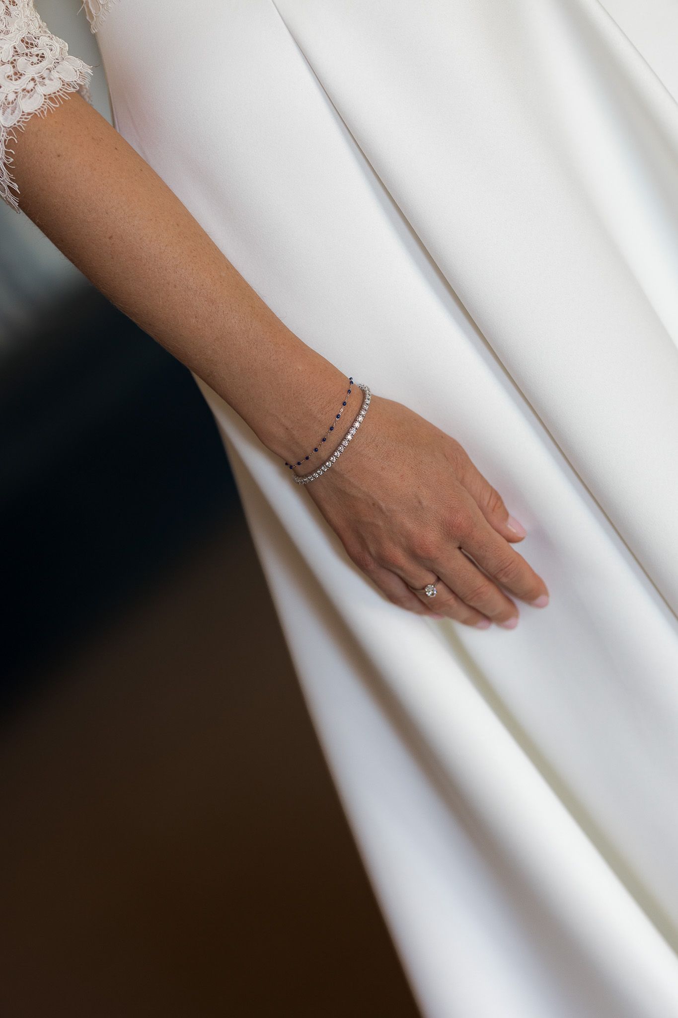 Close-up of a bride's hand in a white wedding dress, wearing a diamond ring and elegant tennis bracelet.