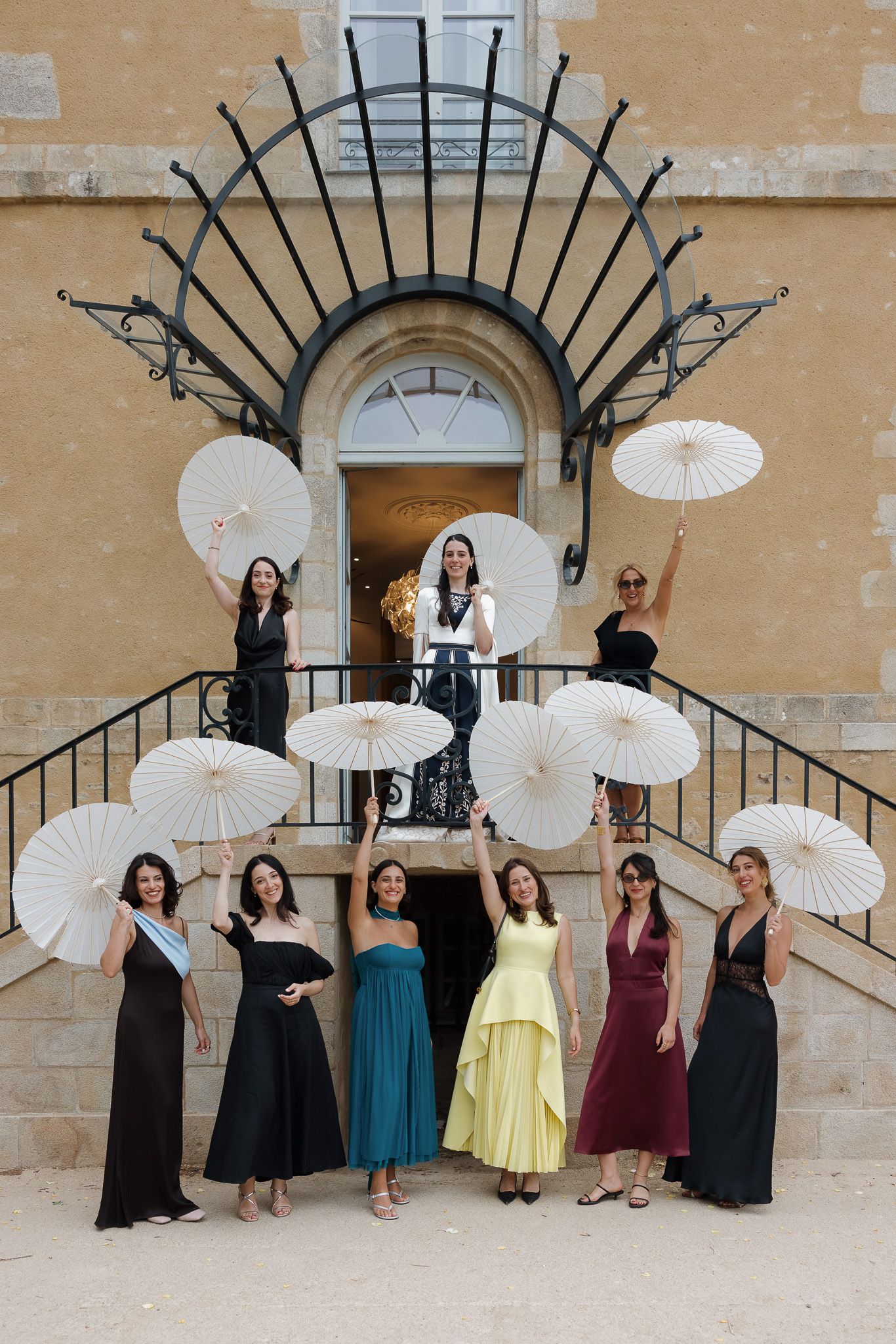 Group of elegant women in colorful gowns holding white parasols on ornate stone staircase at formal event.