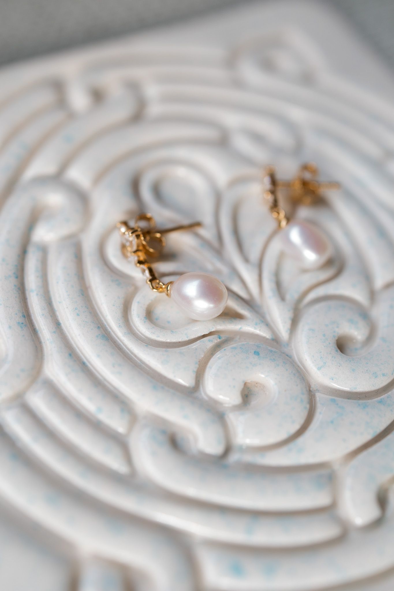 Gold and pearl drop earrings resting on a white decorative ceramic tile with ornate scroll patterns.
