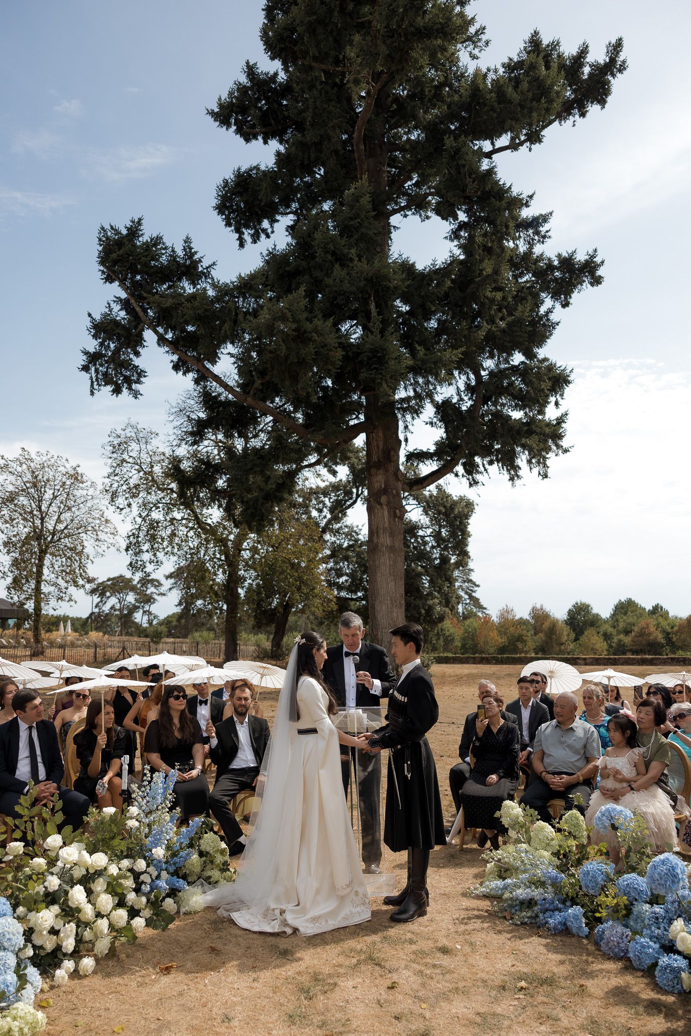 Outdoor wedding ceremony with bride and groom exchanging vows, flanked by blue hydrangeas, guests seated, tall evergreen tree backdrop.