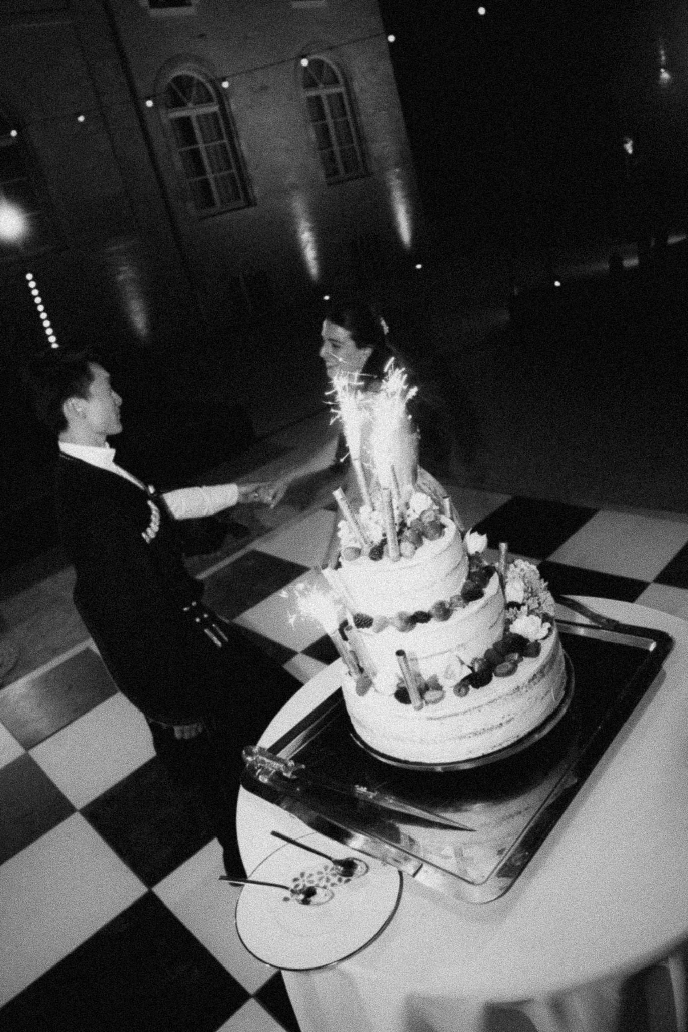Black and white photo of a couple cutting a multi-tiered wedding cake with sparklers on a checkered dance floor.