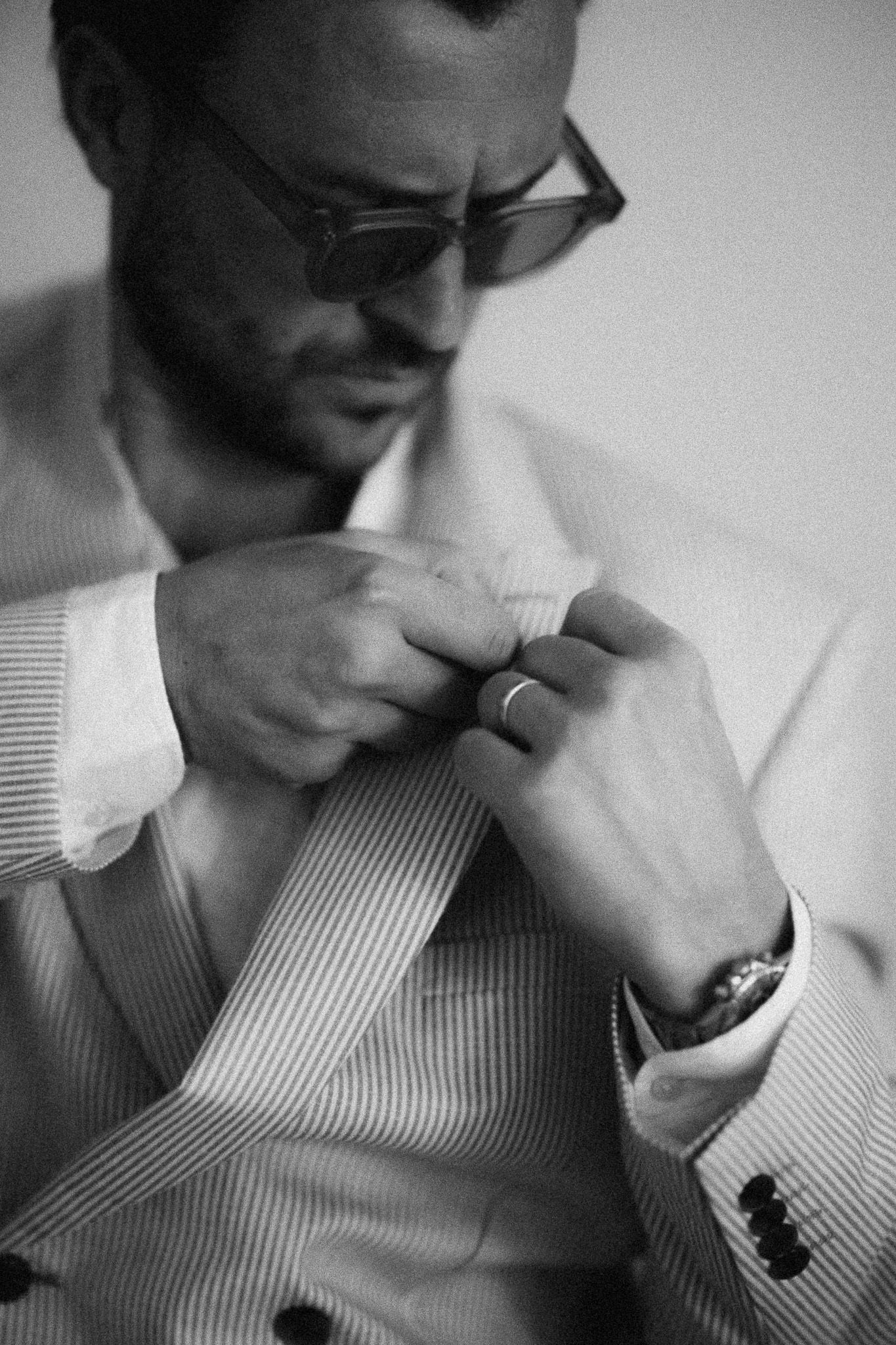 Black and white photo of a well-dressed man in a striped suit adjusting his jacket button, wearing sunglasses and a ring.