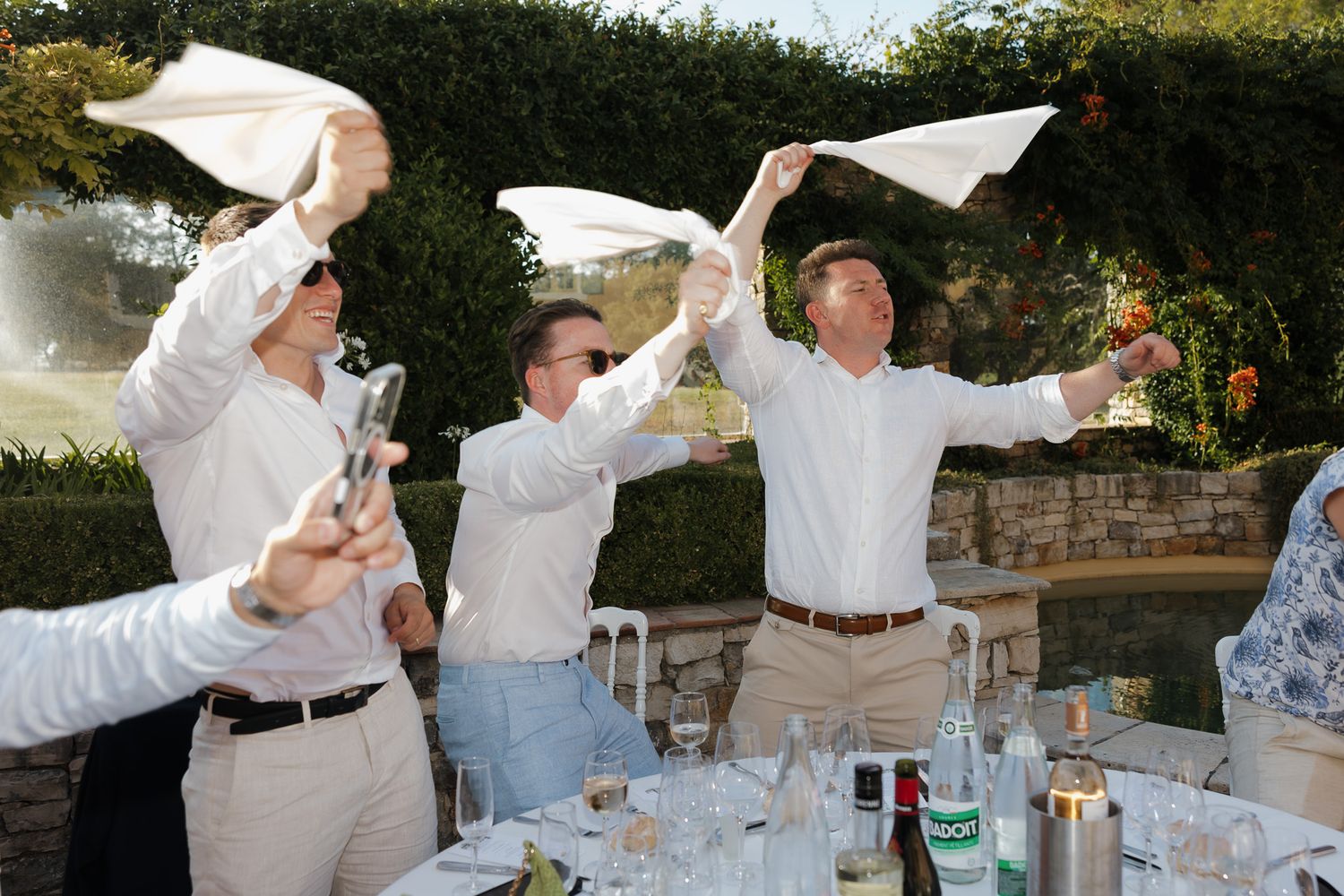 Guests in white shirts wave napkins at an outdoor dinner party with lush garden backdrop.