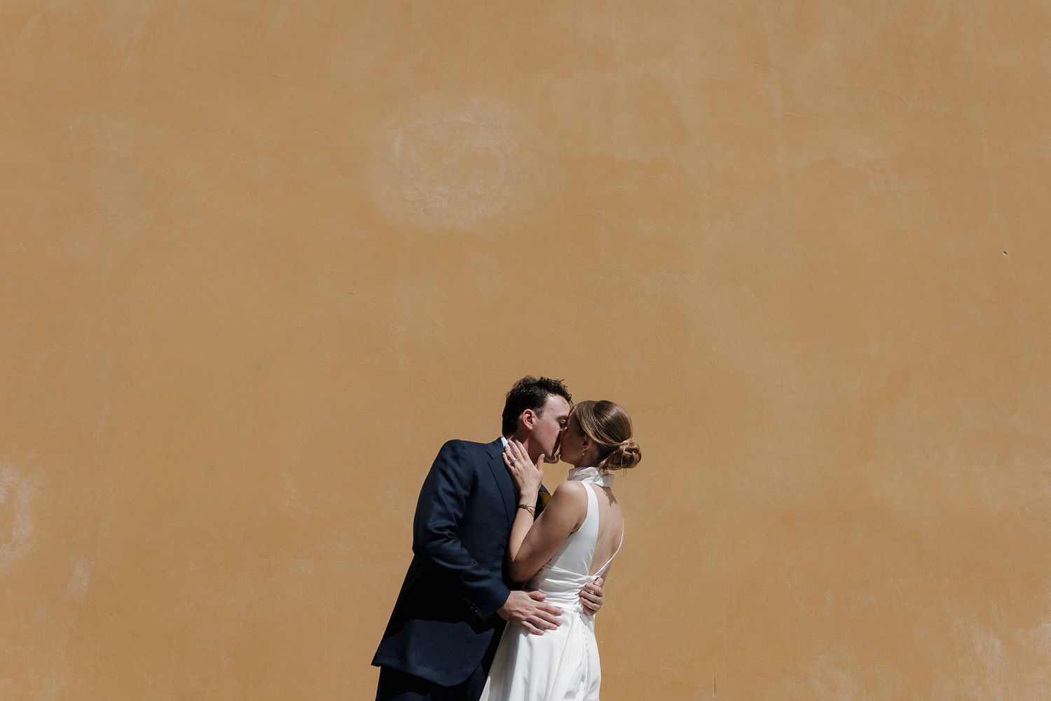 A romantic wedding couple embracing against a warm terracotta colored wall.