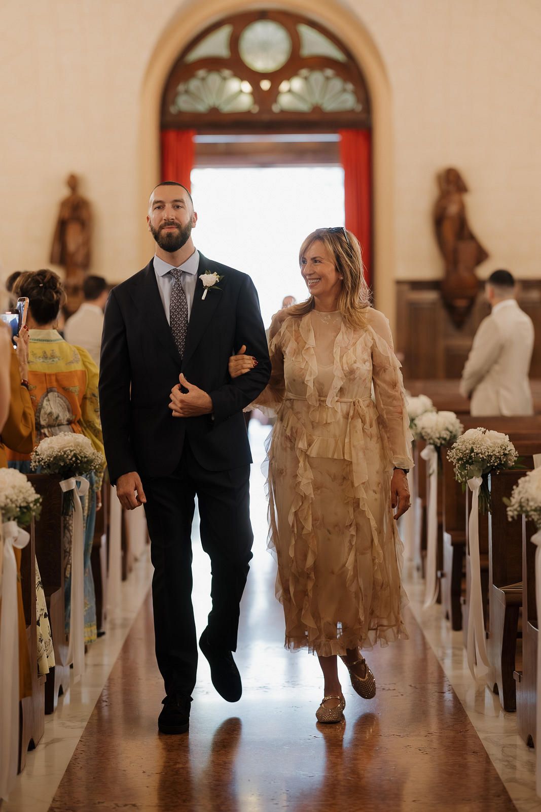 A couple walks down a church aisle decorated with white flowers, with the bride wearing a beige lace dress.