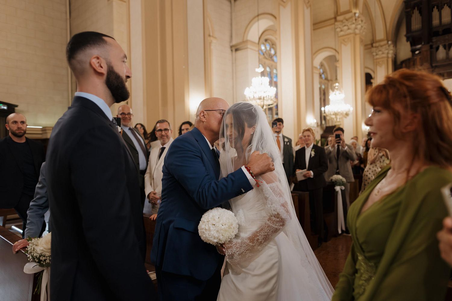 Wedding party gathers in an elegant ballroom with ornate chandeliers and cream-colored walls during a reception celebration.