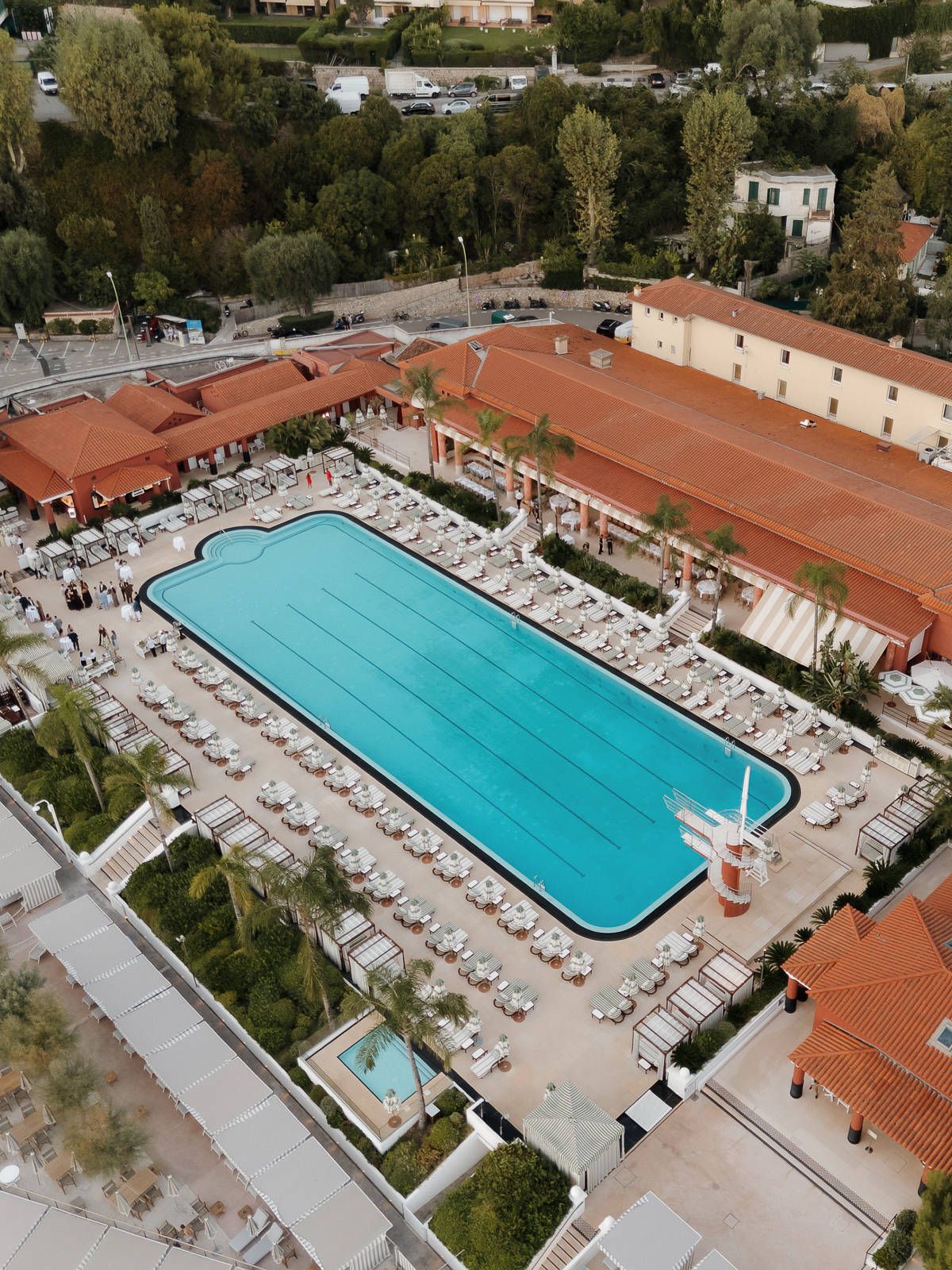 Large rectangular swimming pool surrounded by lounge chairs and terra cotta roofed buildings viewed from above.