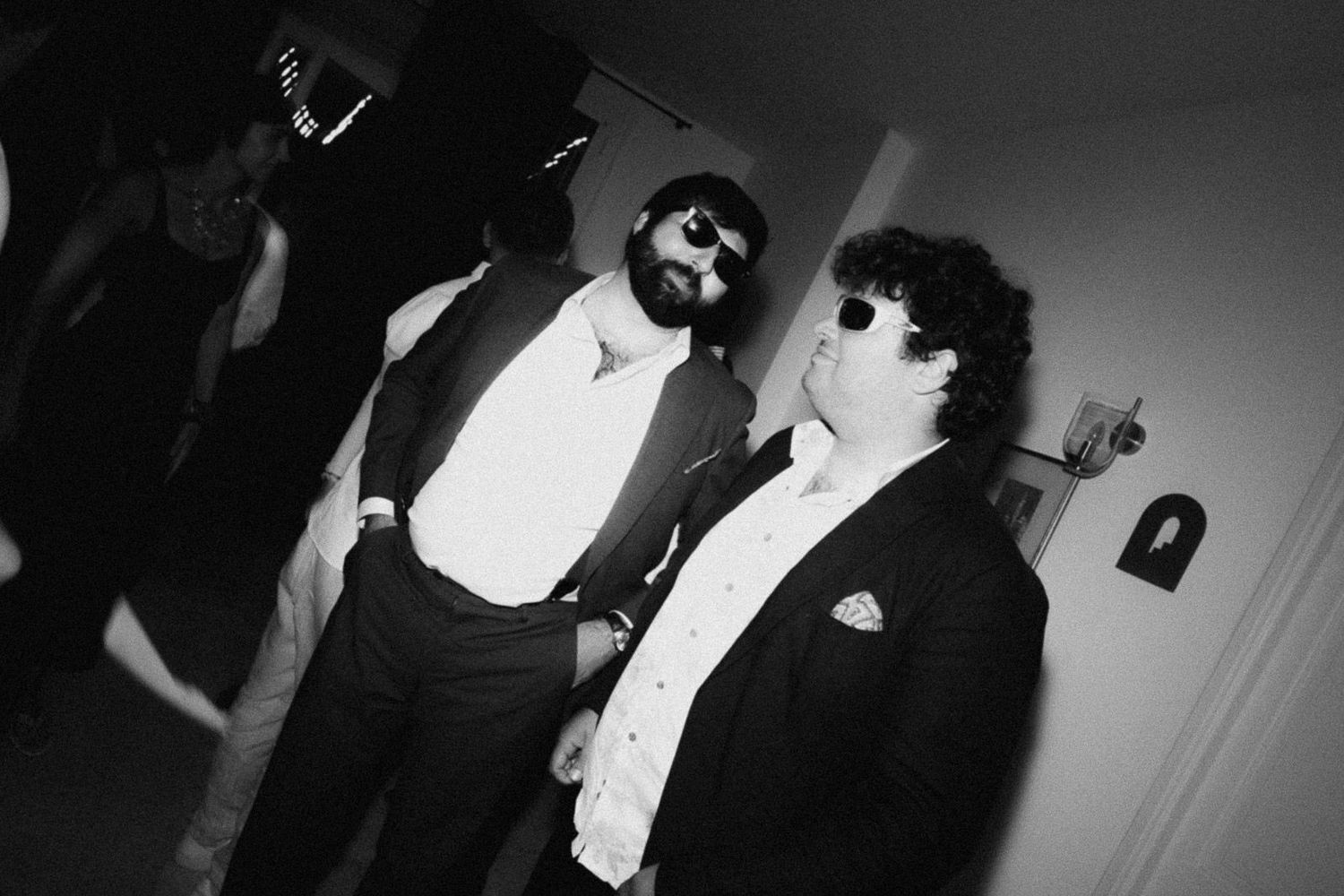 A black and white party photo showing two people in formal attire with tuxedos and bow ties at a social gathering.