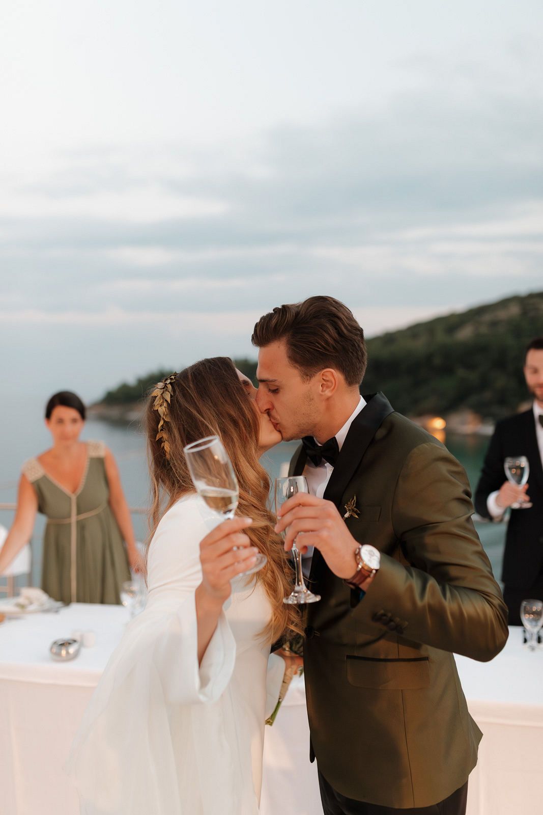 Newlyweds share a romantic kiss while holding champagne glasses at an outdoor coastal wedding reception.
