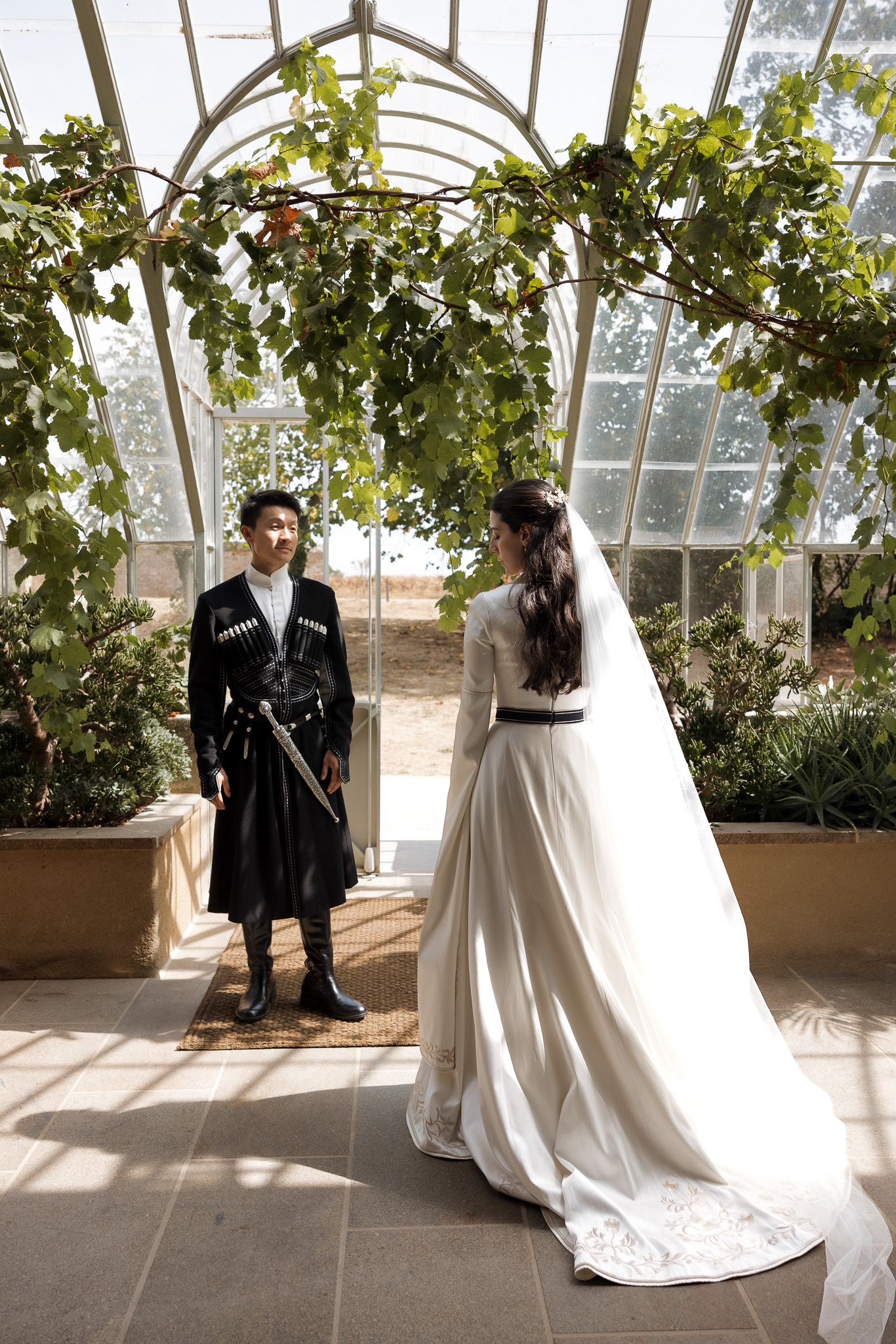 Bride in white gown and groom in traditional black attire stand under green vine arch in sunlit greenhouse.