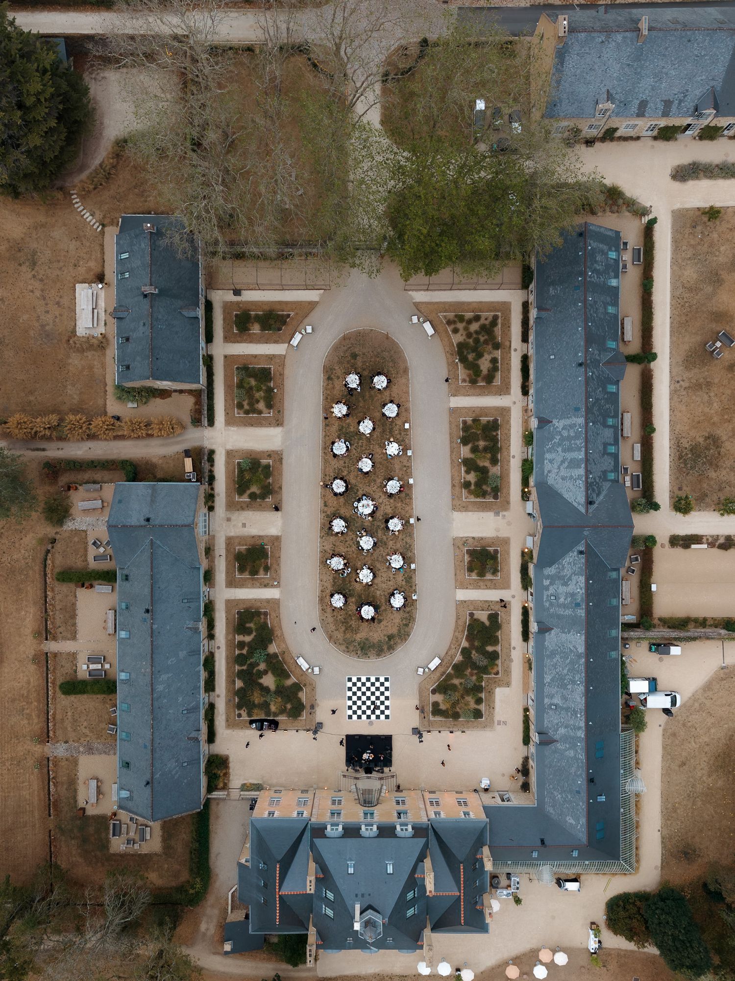 Aerial drone view of a symmetrical estate courtyard with oval garden, white umbrellas, and flanking wings.