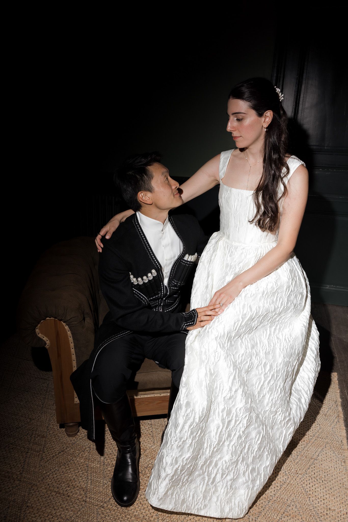 A bride in a white gown and groom in black suit share an intimate moment seated together in dim lighting.