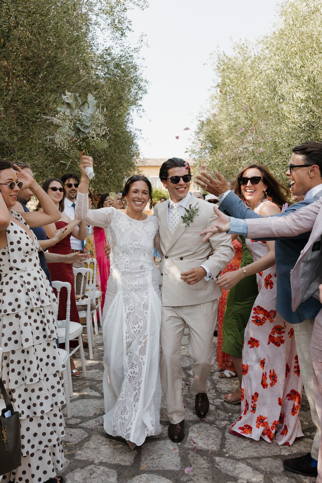 Bride and groom walk through cheering guests outdoors among olive trees during sunny wedding celebration.