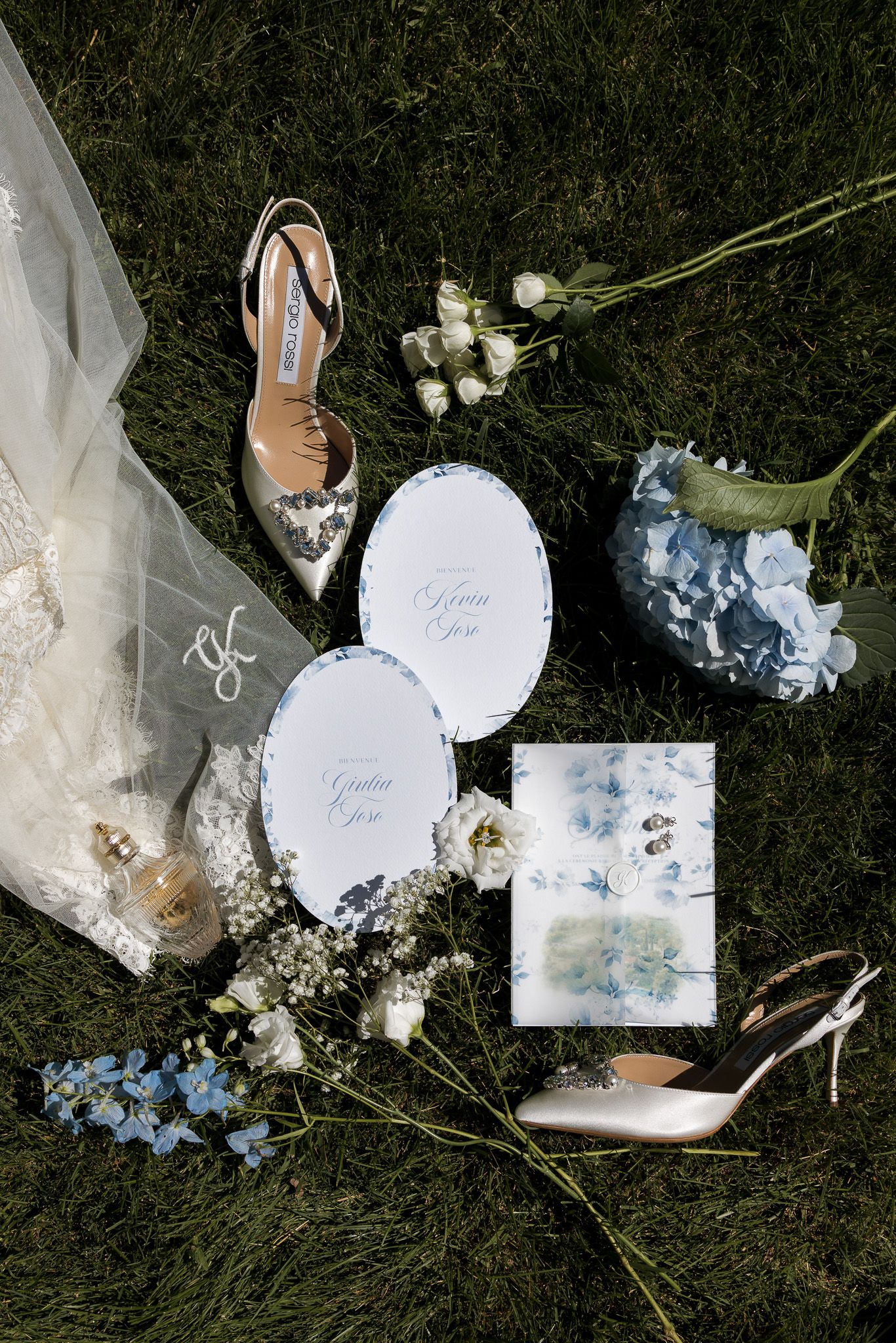Elegant wedding flat lay on grass featuring bridal shoes, blue hydrangeas, white flowers, stationery, and lace dress details.