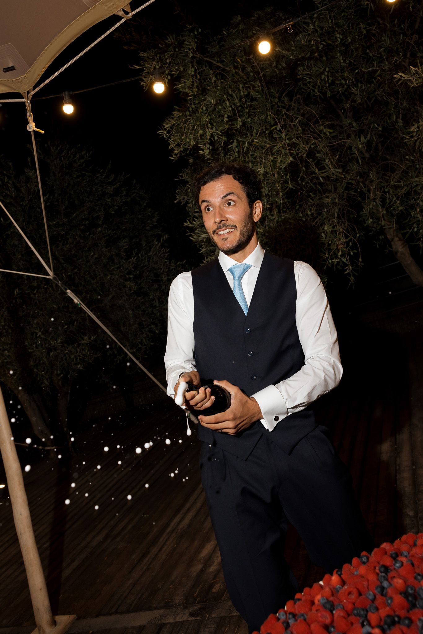 Elegant man in navy vest and blue tie opening champagne bottle at outdoor evening event with string lights.