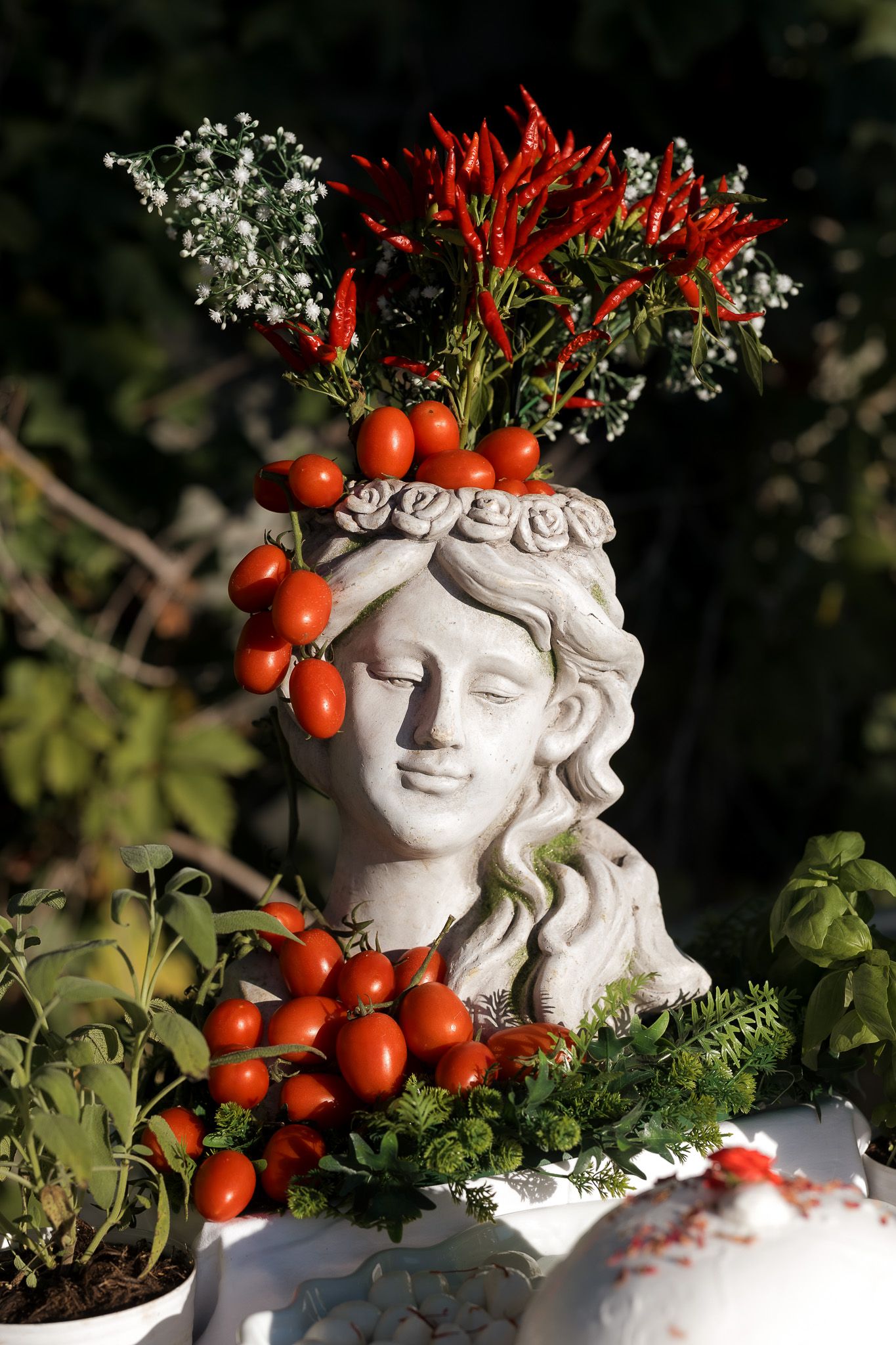 Stone bust of a woman decorated with cherry tomatoes, red chili peppers, and fresh herbs outdoors.