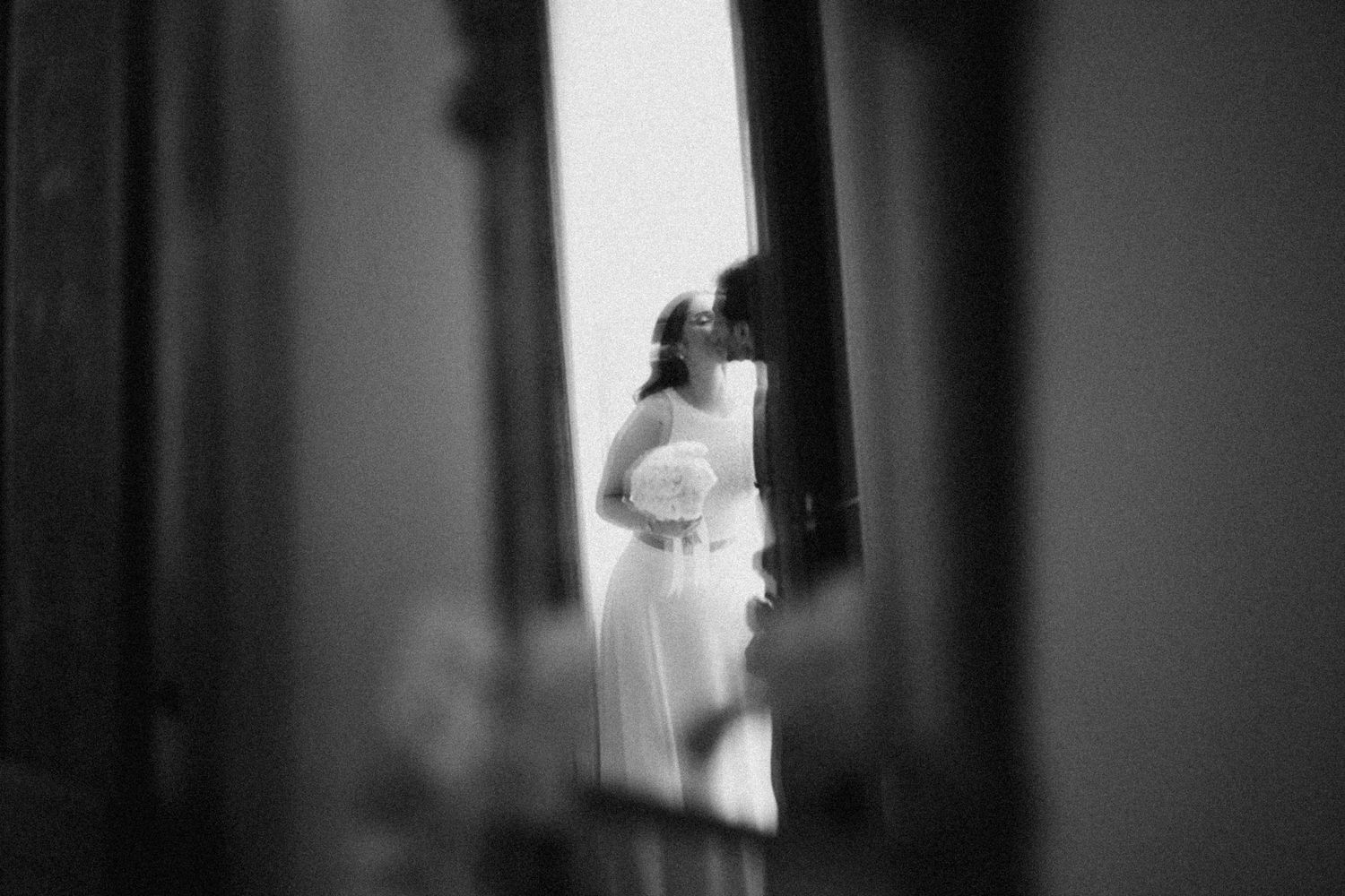 Black and white wedding photo of bride in white gown reflected in mirror, holding bouquet.