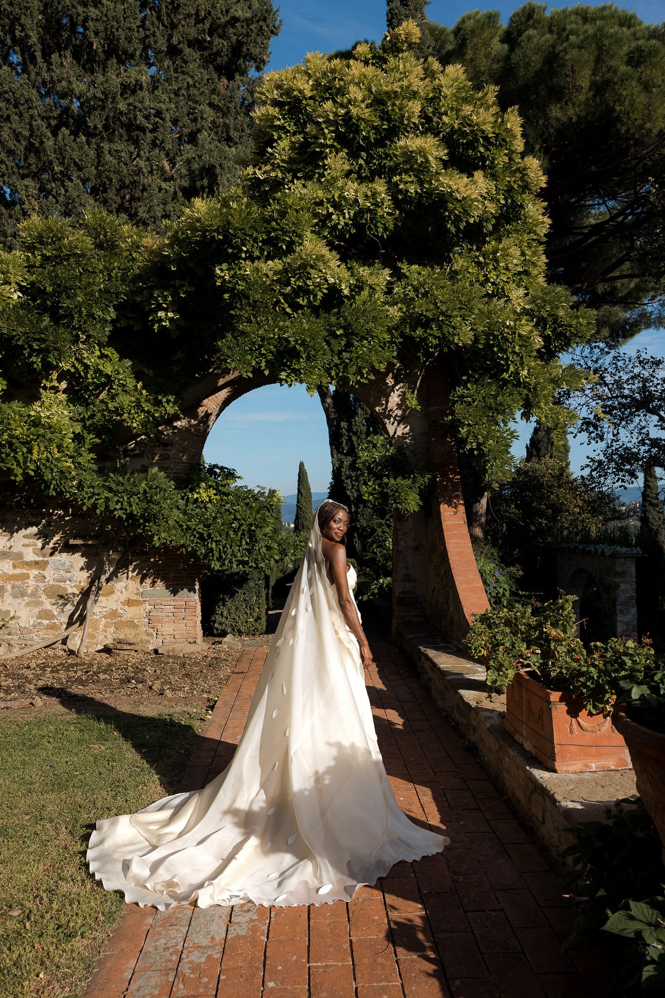 Bride in a flowing white wedding gown with a long train stands on a brick path beneath a vine-covered stone arch in a sunlit garden.