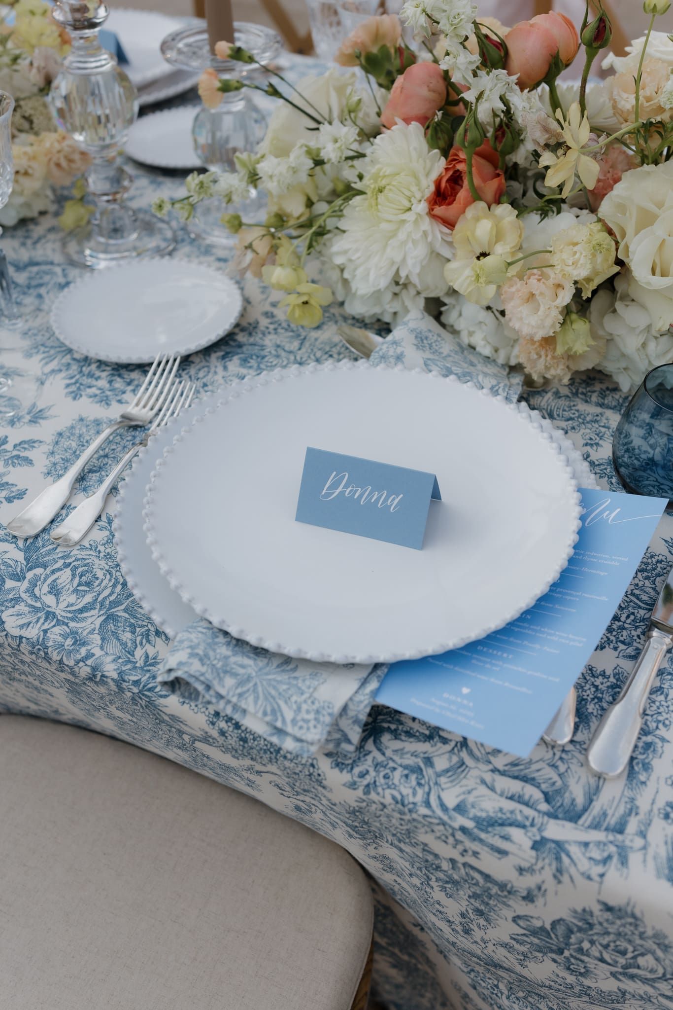 Elegant table setting with white scalloped plates on light blue toile placemats with soft floral arrangements.