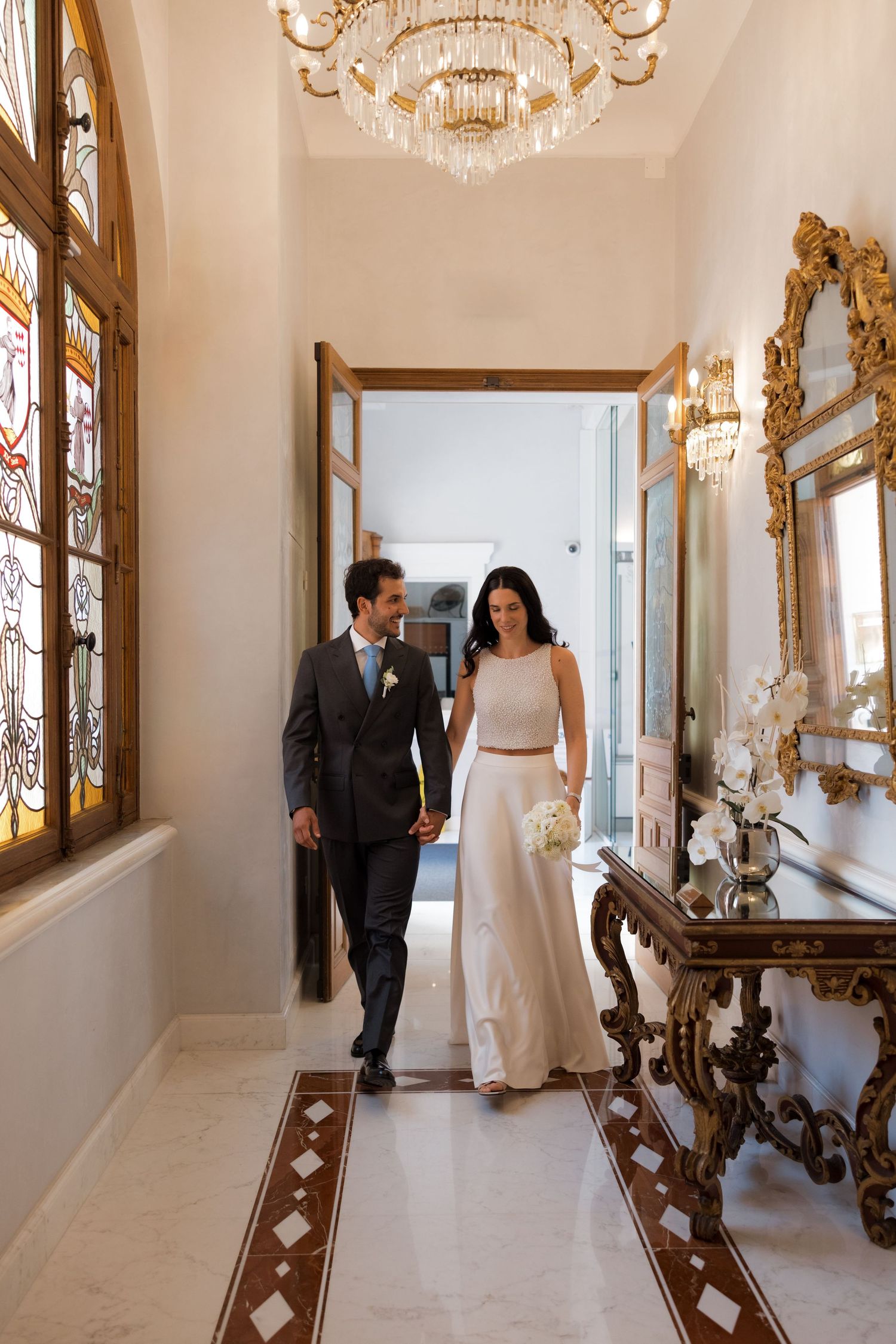 Elegant bride and groom walking hand in hand through ornate hallway with chandelier and stained glass windows.