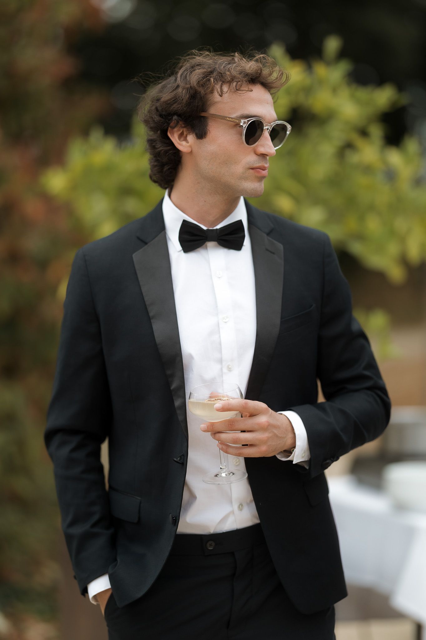Elegant man in black tuxedo with bow tie and sunglasses holding a champagne glass outdoors.