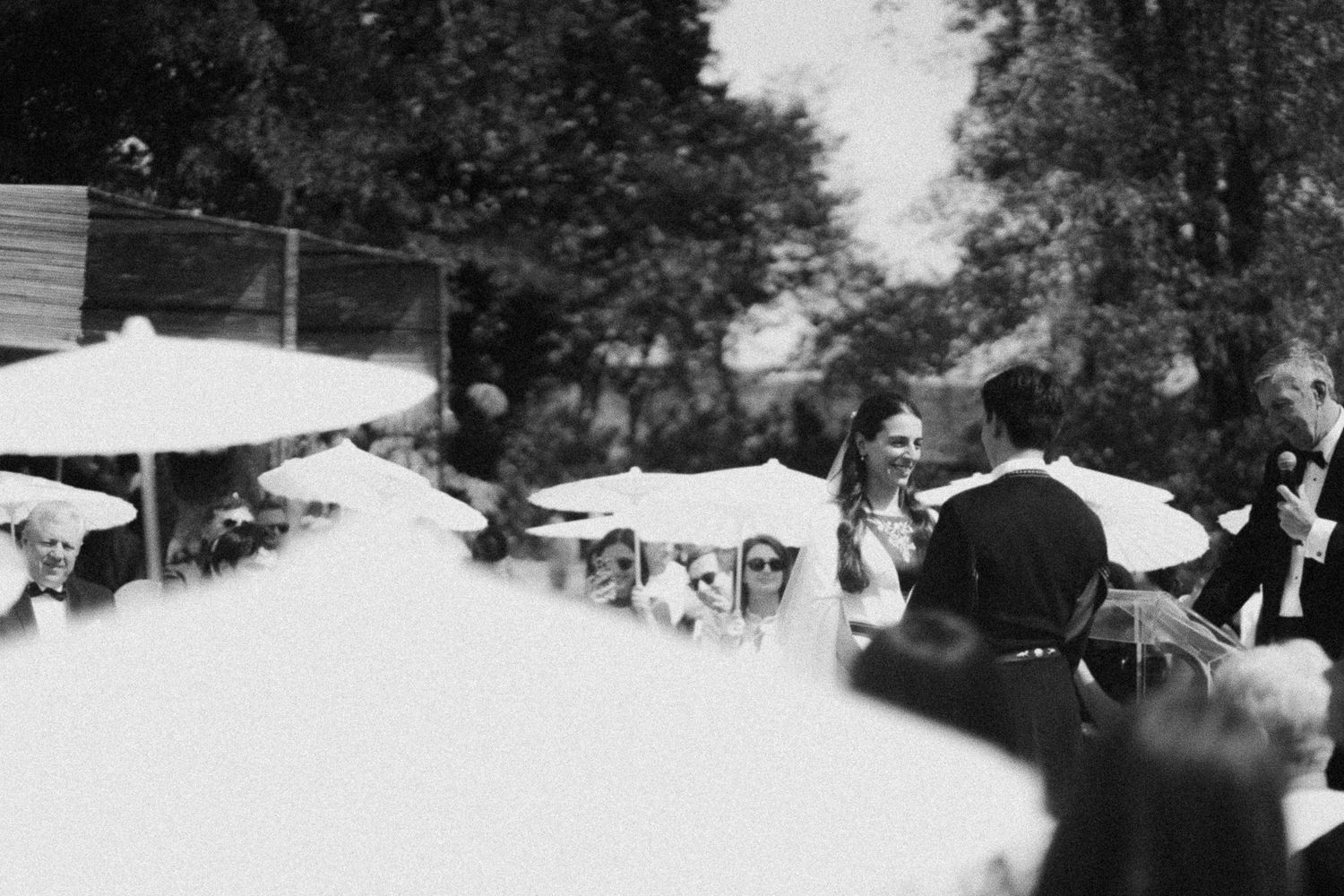 Black and white outdoor wedding ceremony with bride, groom, and guests under white umbrellas amid lush trees.