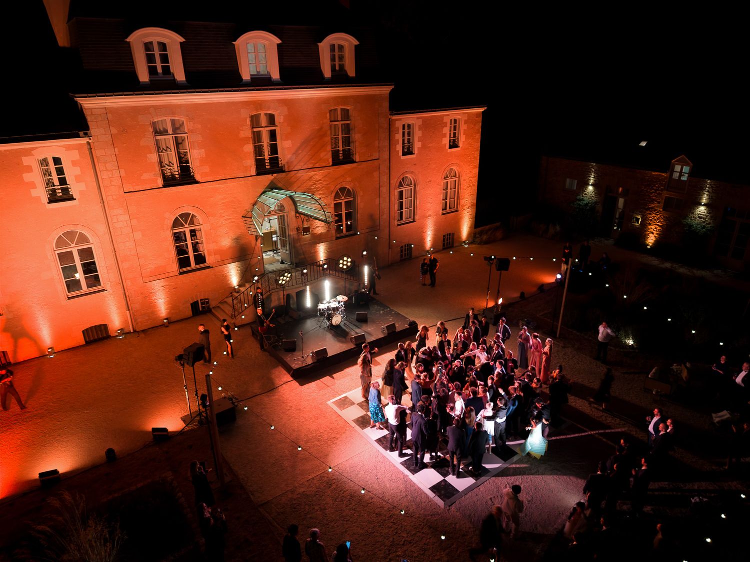 Aerial night view of elegant French chateau courtyard with guests gathered at an outdoor evening event, illuminated by warm orange lighting.