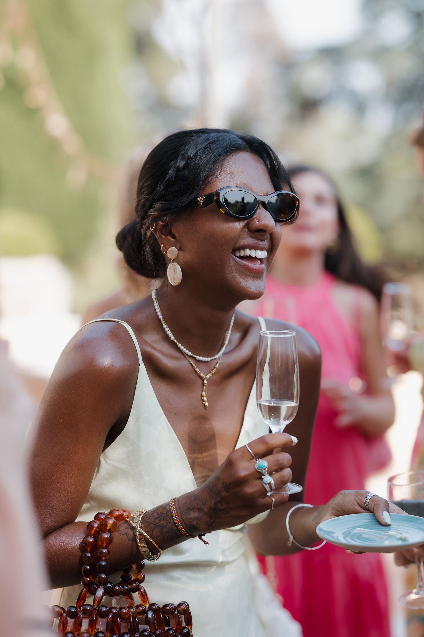 A smiling woman in sunglasses holds a champagne flute at an outdoor party, wearing a gold dress and layered necklaces.