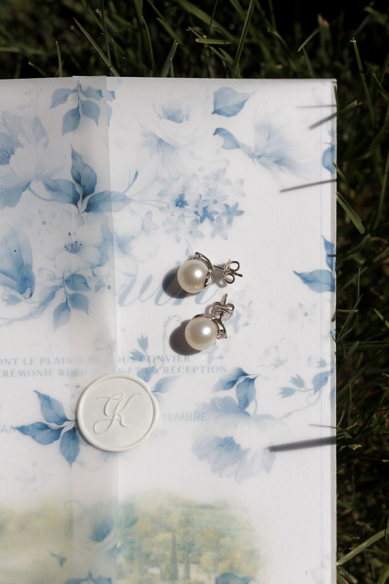 Pearl drop earrings on floral blue wedding stationery with wax seal initial K, resting on green grass.