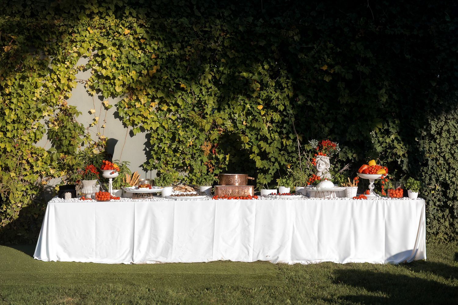 Elegant outdoor buffet table with white linen and colorful vegetable arrangements against ivy-covered wall.