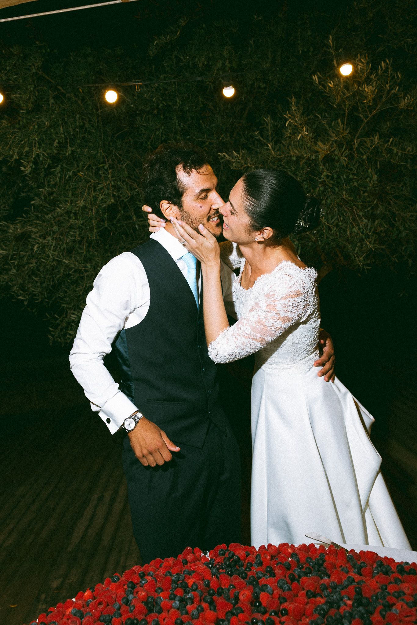 A bride and groom share a romantic kiss surrounded by red flowers and string lights at their outdoor wedding reception.