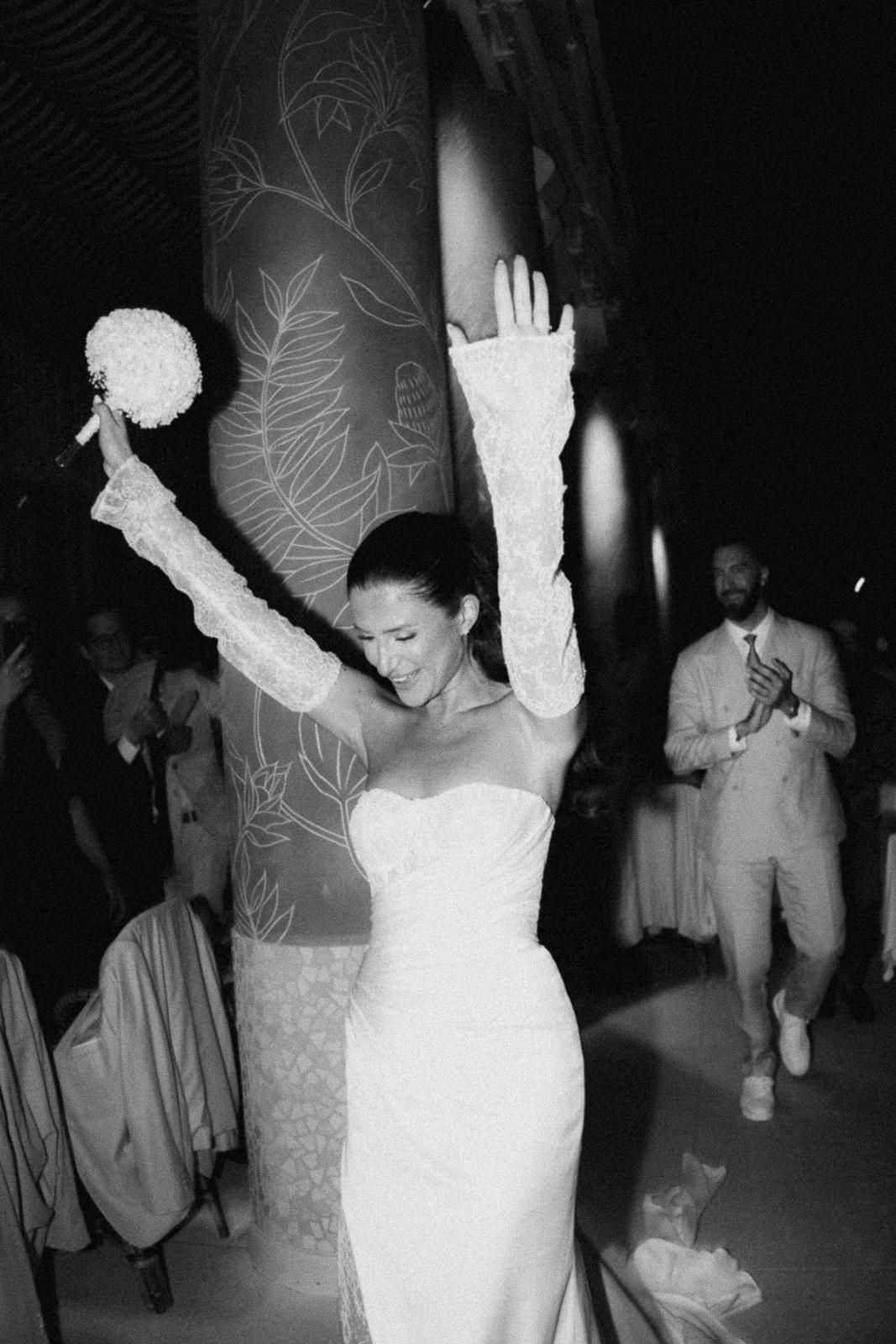 A bride in a fitted white lace gown dances joyfully with raised arms at a wedding reception.