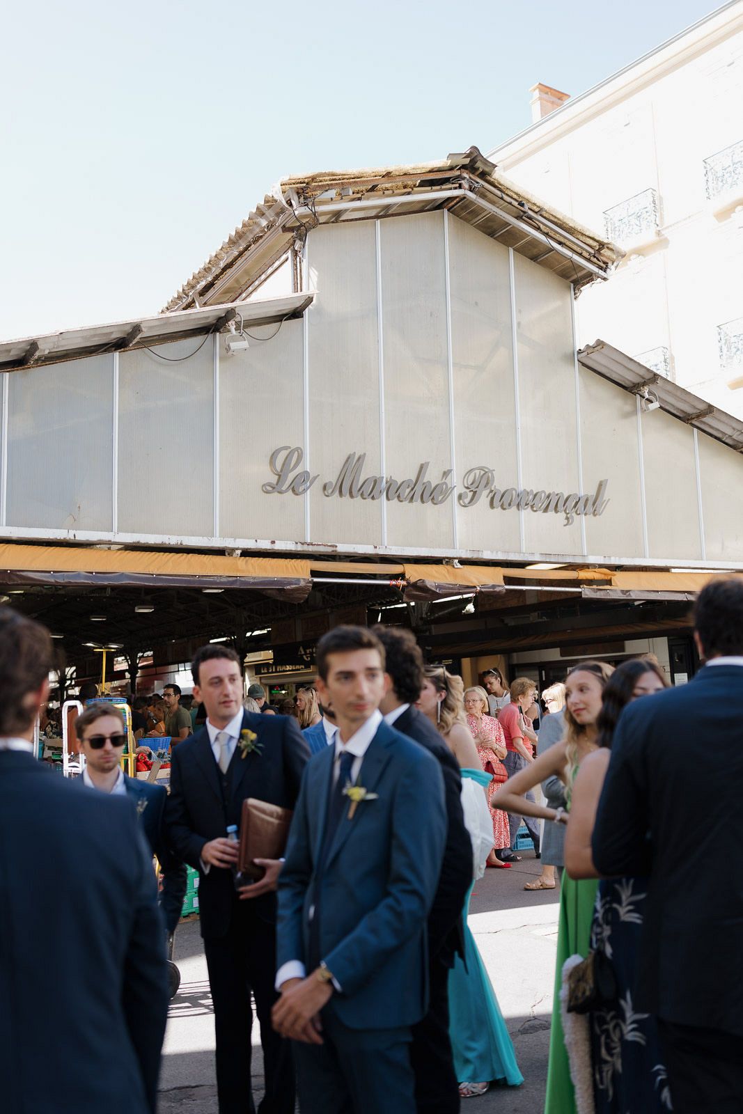 Guests in formal attire gather at an outdoor venue with white canopy structures visible in the background.