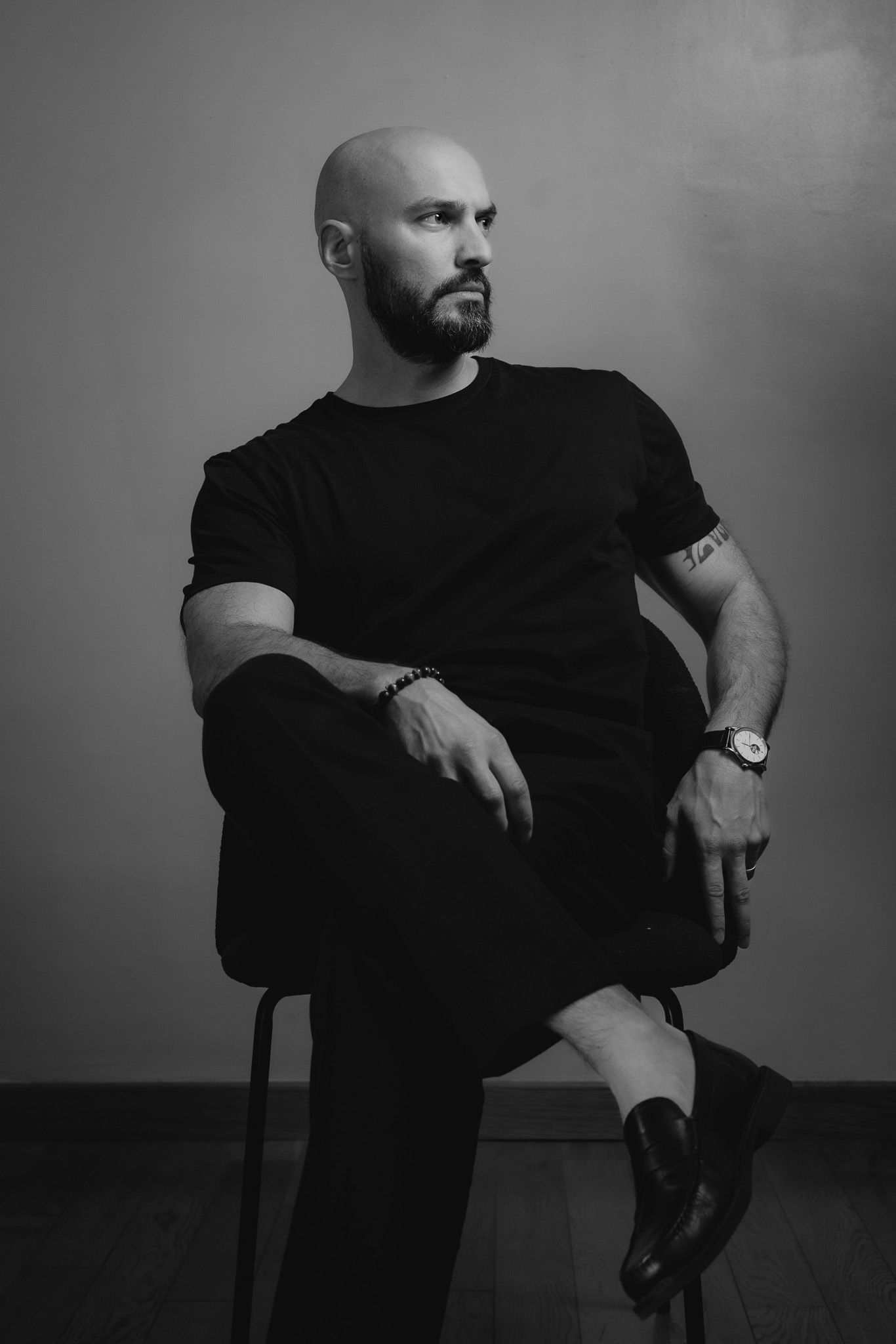 Black and white portrait of a bald, bearded man in a black t-shirt and trousers sitting cross-legged on a chair, looking to the side.