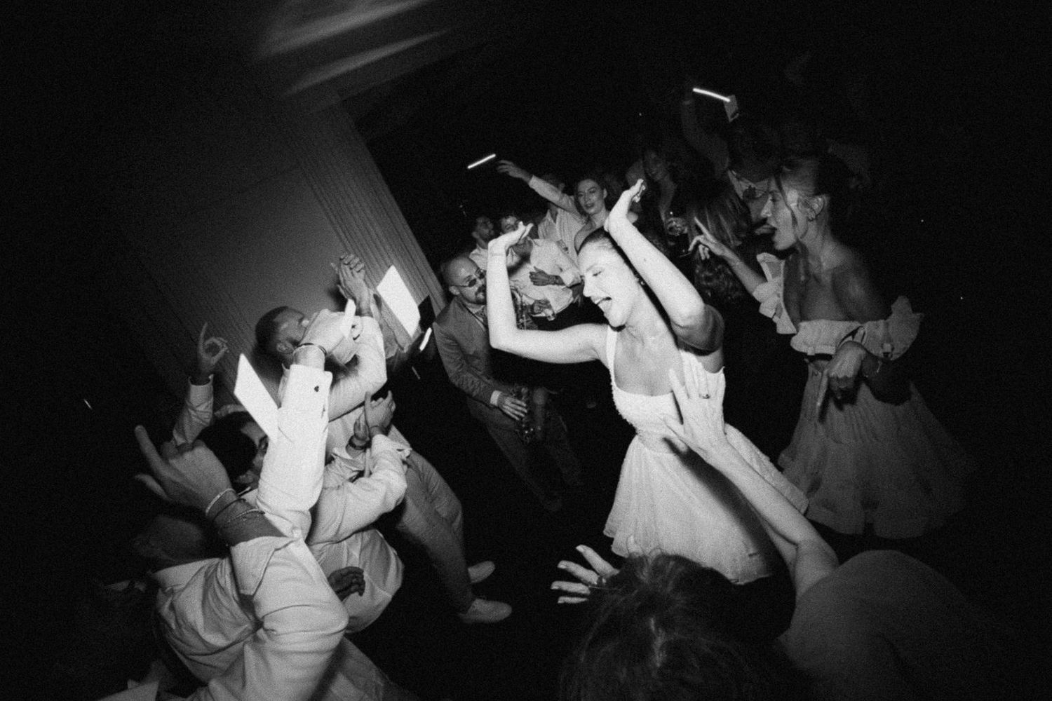 Black and white photo of people dancing energetically at a party with raised arms and dynamic movement.