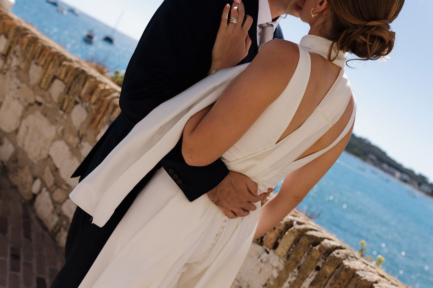 A couple in elegant wedding attire embraces on a scenic balcony overlooking a bright blue Mediterranean coastline.